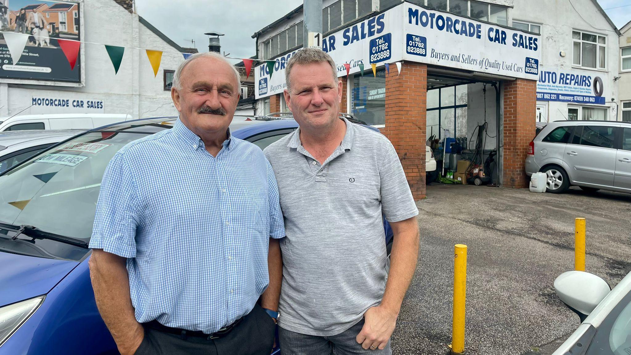 Two men stand in front of a car garage and dealership.