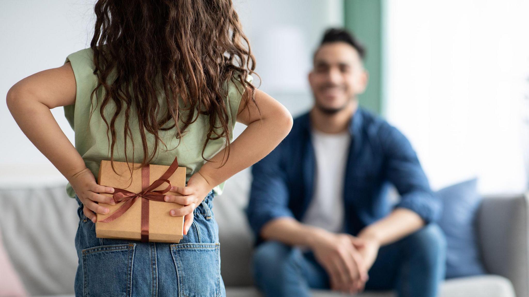 a girl facing her dad who is out of focus, the girl is holding a present wrapped in brown paper with a brown ribbon on it behind her back.