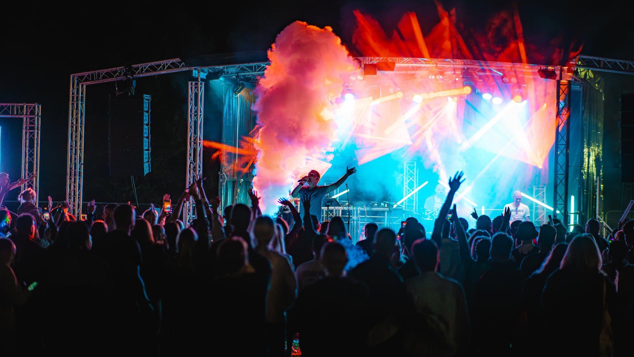A dark image taken from behind a large audience showing a band performing on stage. There are colourful lights on the stage and smoke.