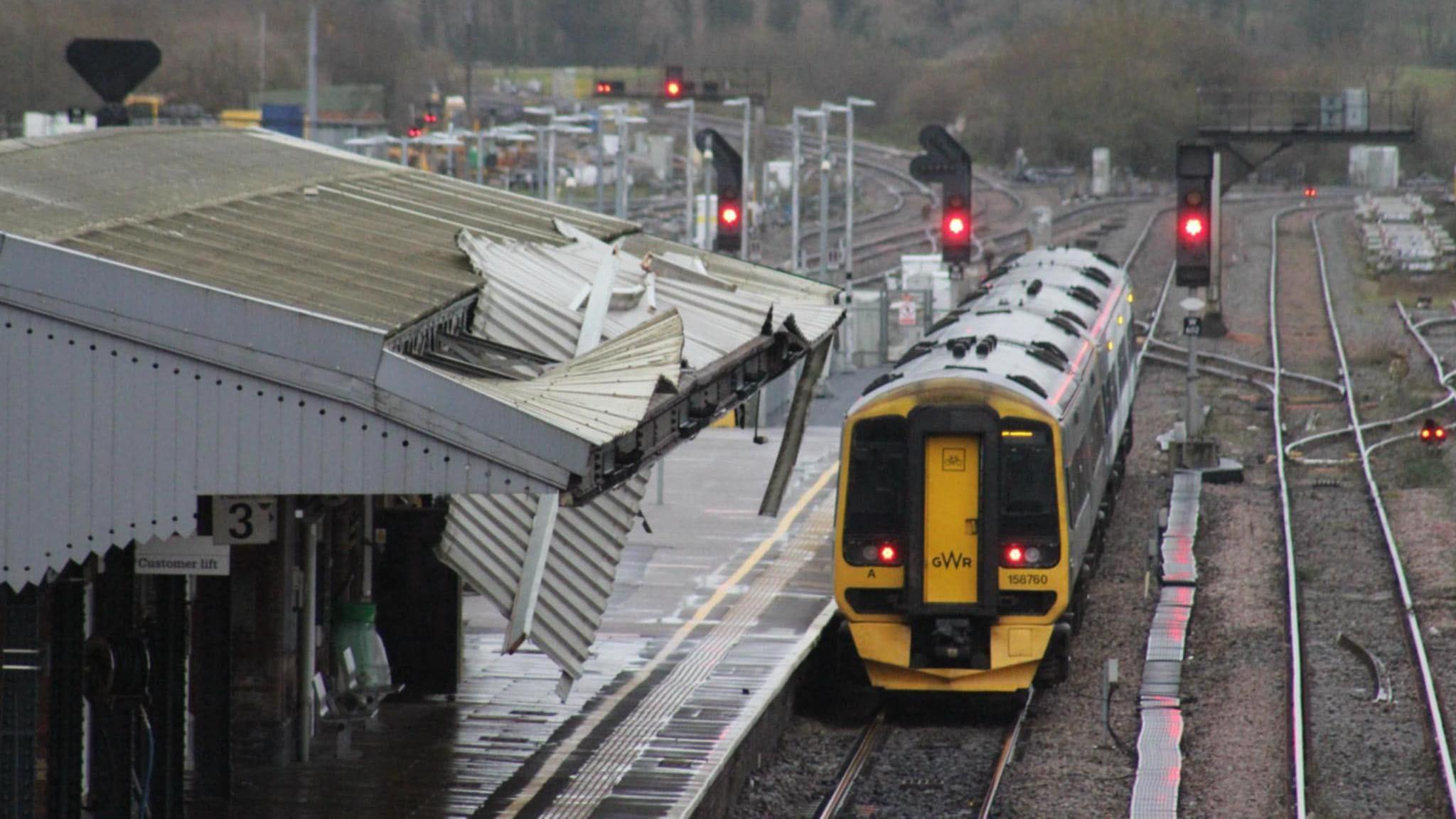 A train pulled up at the platform at Westbury station. It is a yellow and green GWR train. A canopy over the platform edge can be seen, but it is damaged from Storm Darragh. Parts of the roof are hanging down and gaping holes can be seen. 