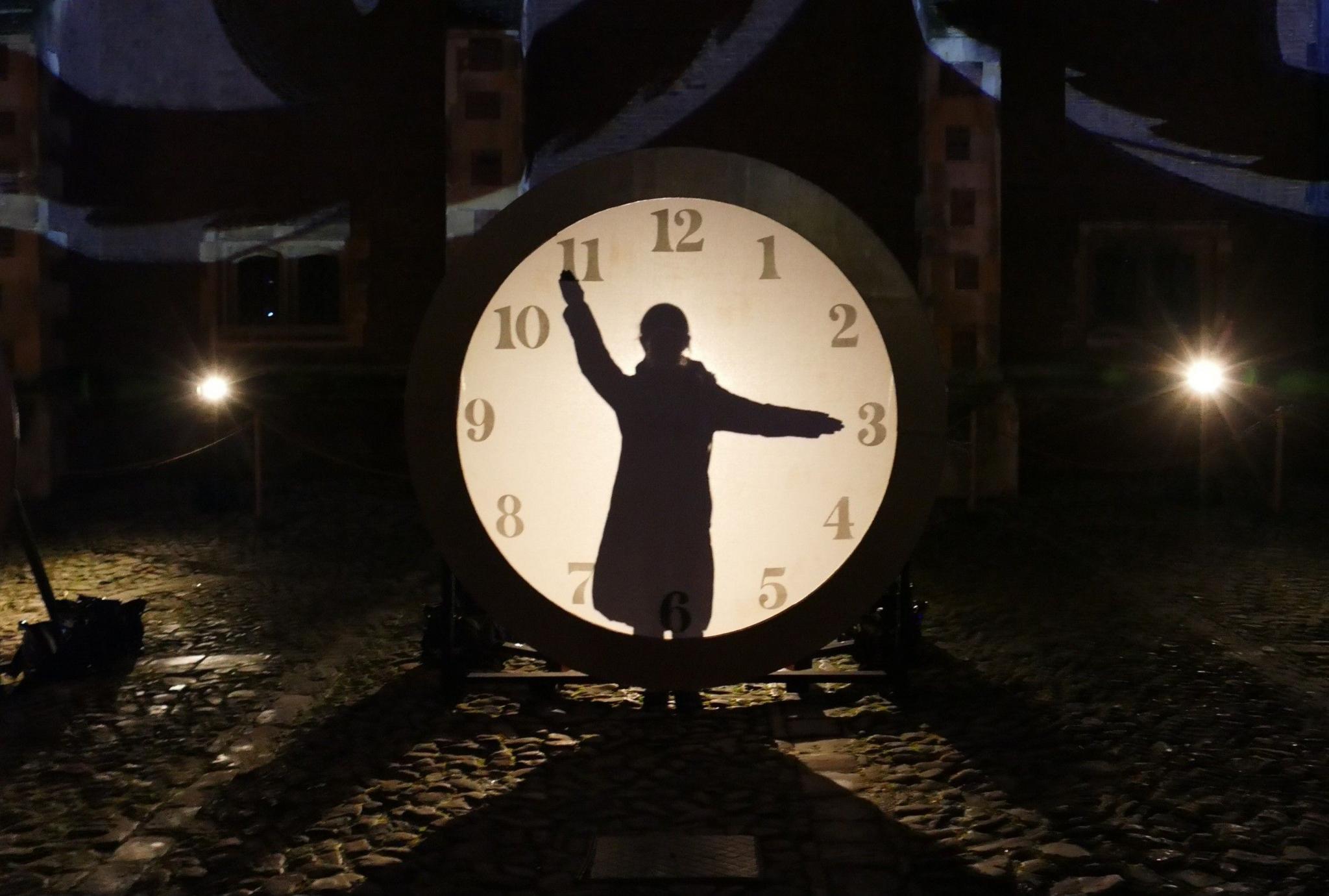 A person is seen in silhouette, standing in front of an illuminated clock, which appears taller than the person