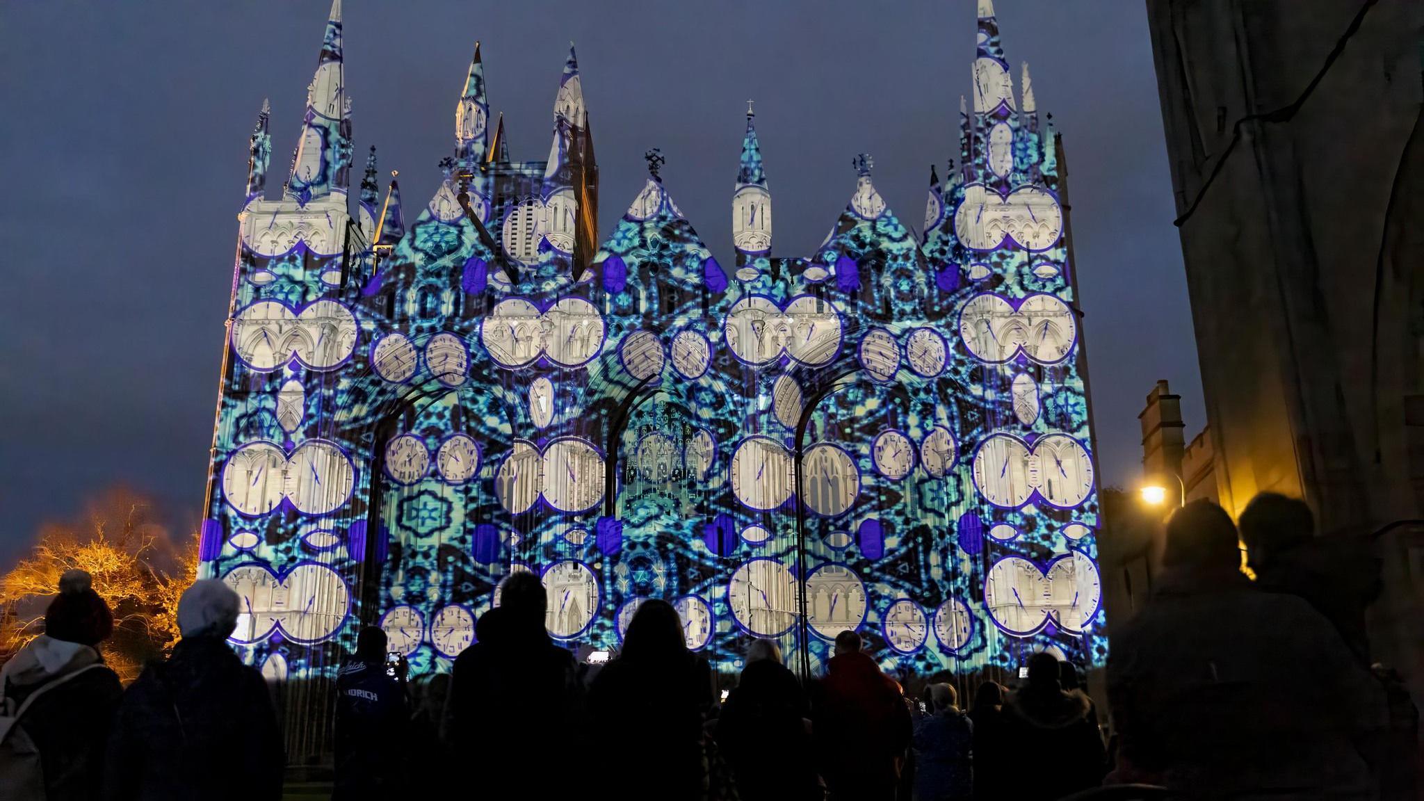 The exterior of Peterborough Cathedral lit up at night with a projection showing many clock faces. In the foreground are people with winter clothing looking at it.