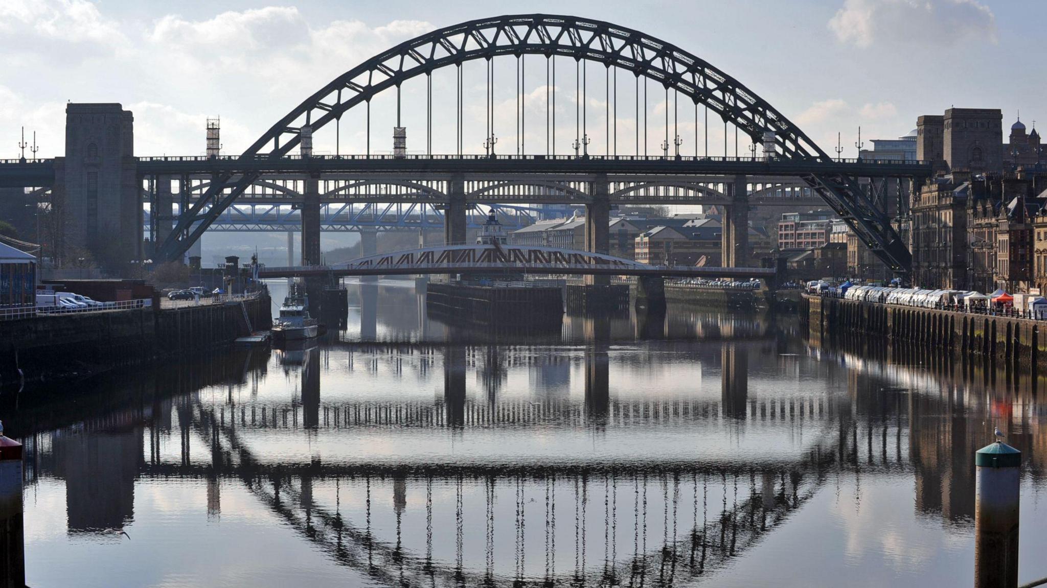 A wide view of the Tyne Bridge from the river Tyne. It is a metal bridge, painted green. Several other bridges can be seen behind it. The silhouettes of the bridges are reflected in the water below.