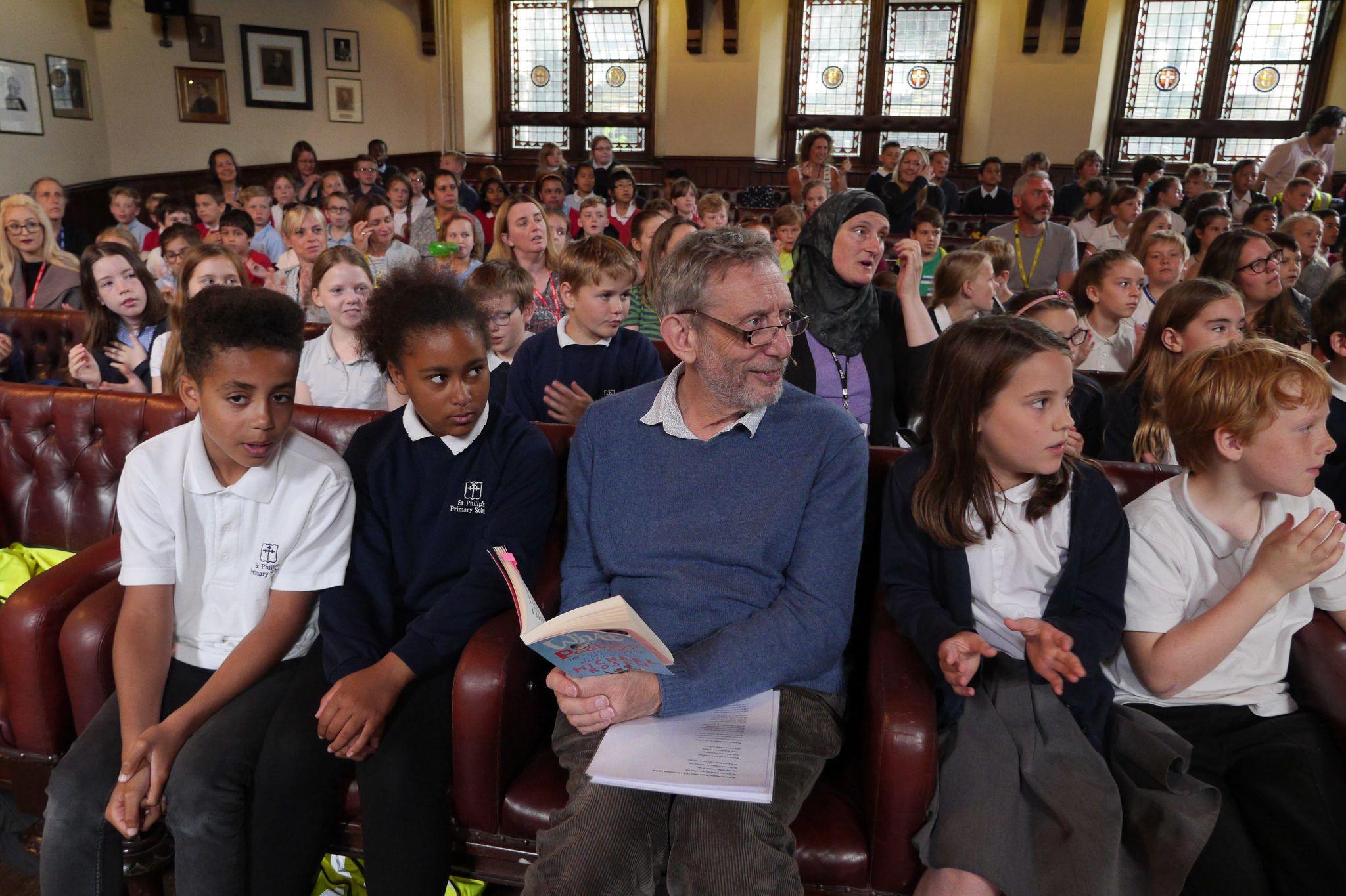 Michael Rosen sits in a hall full of children.