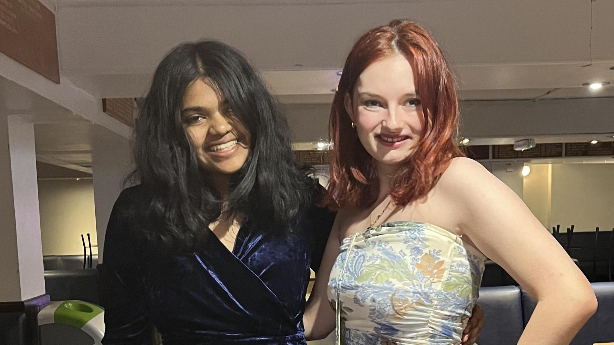 Vanya and Hannah standing together smiling at the camera. The girl on the left has dark hair and is wearing a dark blue, velvet wrap dress. The girl on the right has red-coloured hair and is wearing a cream dress with blue, green and orange floral pattern.