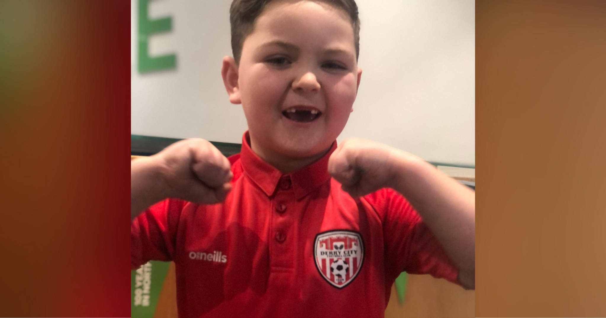 Six-year-old Derry City fan Braelin proudly wears a team polo shirt. He has brown hair and is stranding in front of a row of flags