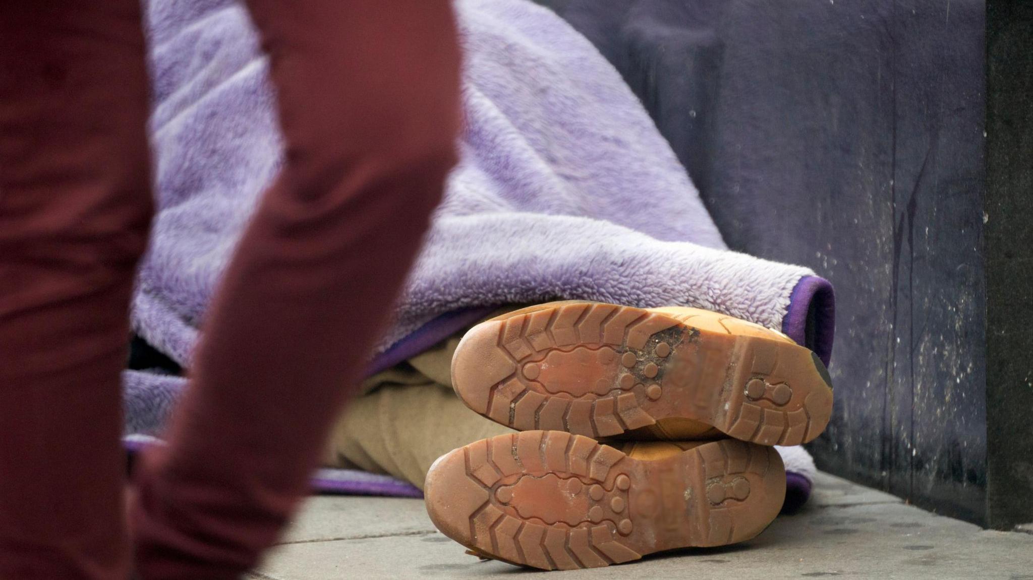 A person lies on the pavement wrapped in a purple blanket, with only their boots visible, as someone walks past in the foreground.