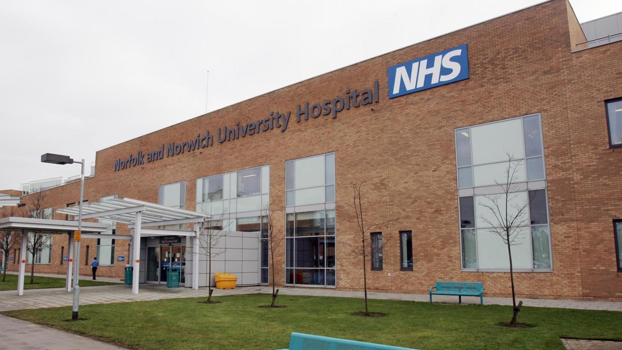 A brown bricked hospital which has large glass windows and a patch of grass with planted trees out the front. A sign on the building reads Norfolk and Norwich University Hospital with an NHS logo next to it.