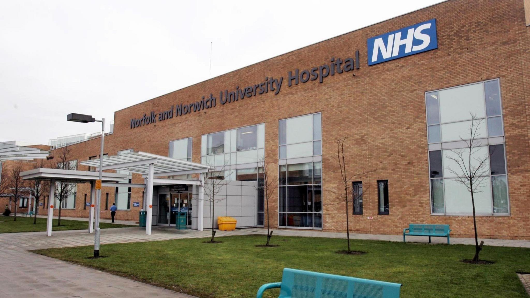 The entrance of the hospital with a sign which reads "Norfolk and Norwich University Hospital NHS". The hospital has brown bricks and large windows