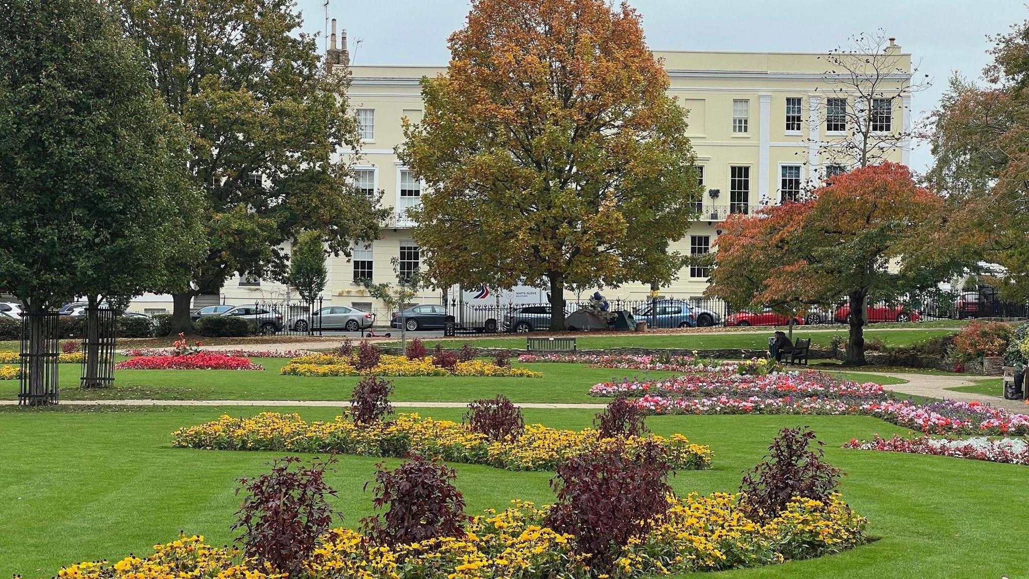 A green park, full of colourful, shaped flowerbeds, trees with turning leaves and winding paths, stands in a square in Cheltenham town centre. Large, Regency homes border the park on the other side of the road, which motorists have parked their vehicles on.