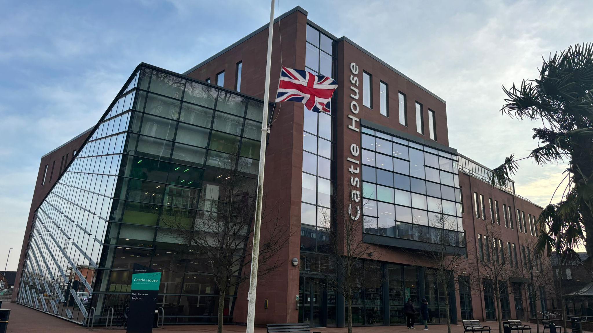 A large brick and glass building bearing the words Castle House. In the foreground, the red, white and blue union flag is flying at half mast.