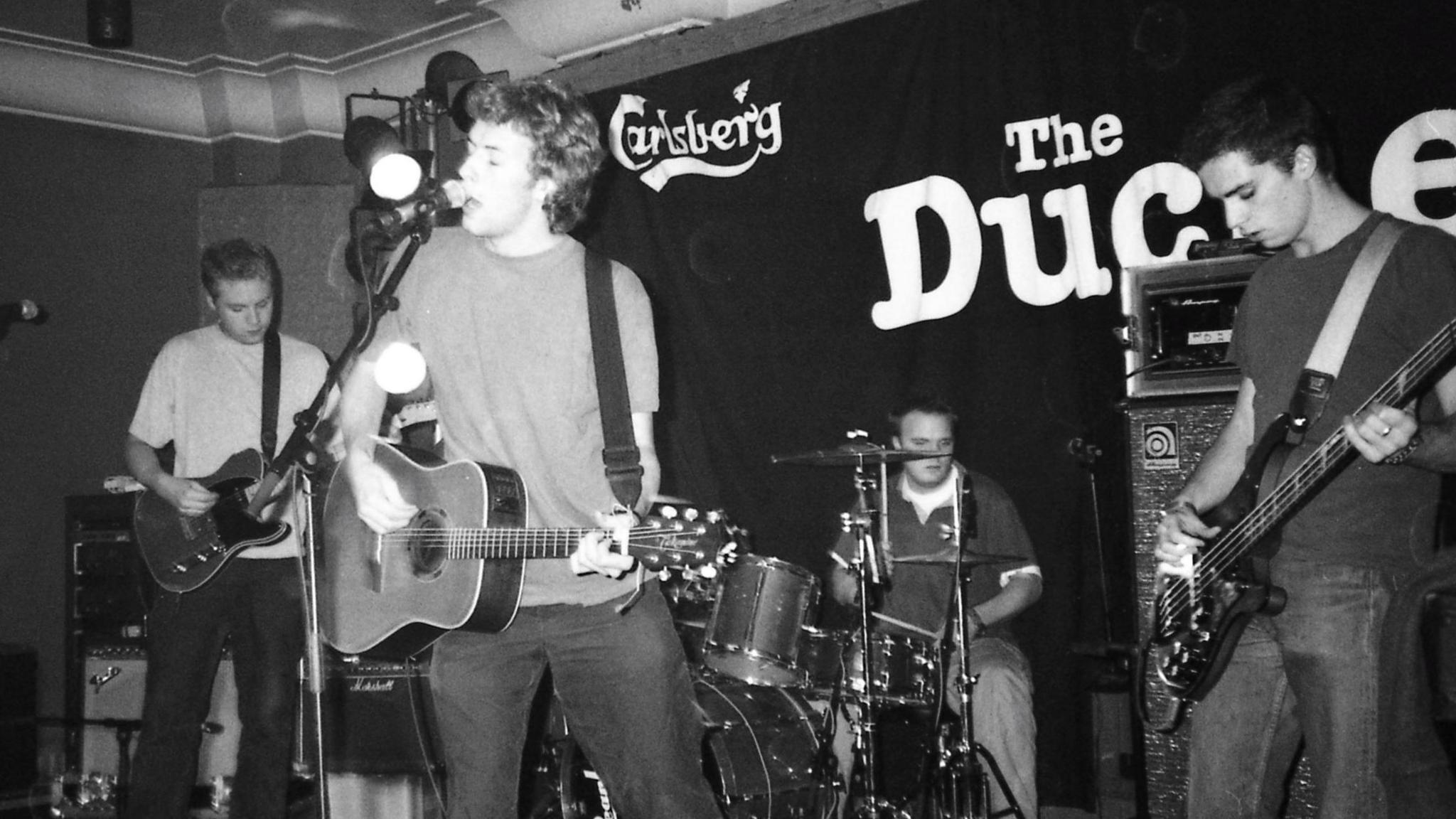 A grainy black and white image of four men playing various musical instruments on a stage. In the background is a black sign with white writing which says Carslberg and The Duchess.