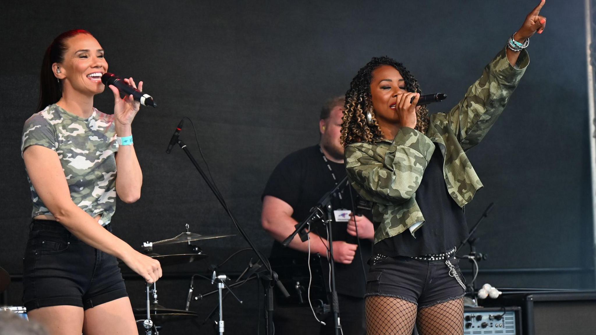 Two members of the band Liberty X, both in black shorts with one wearing a camouflage T-shirt and the other a black T-shirt and camouflage jacket, sing into microphones and dance on stage