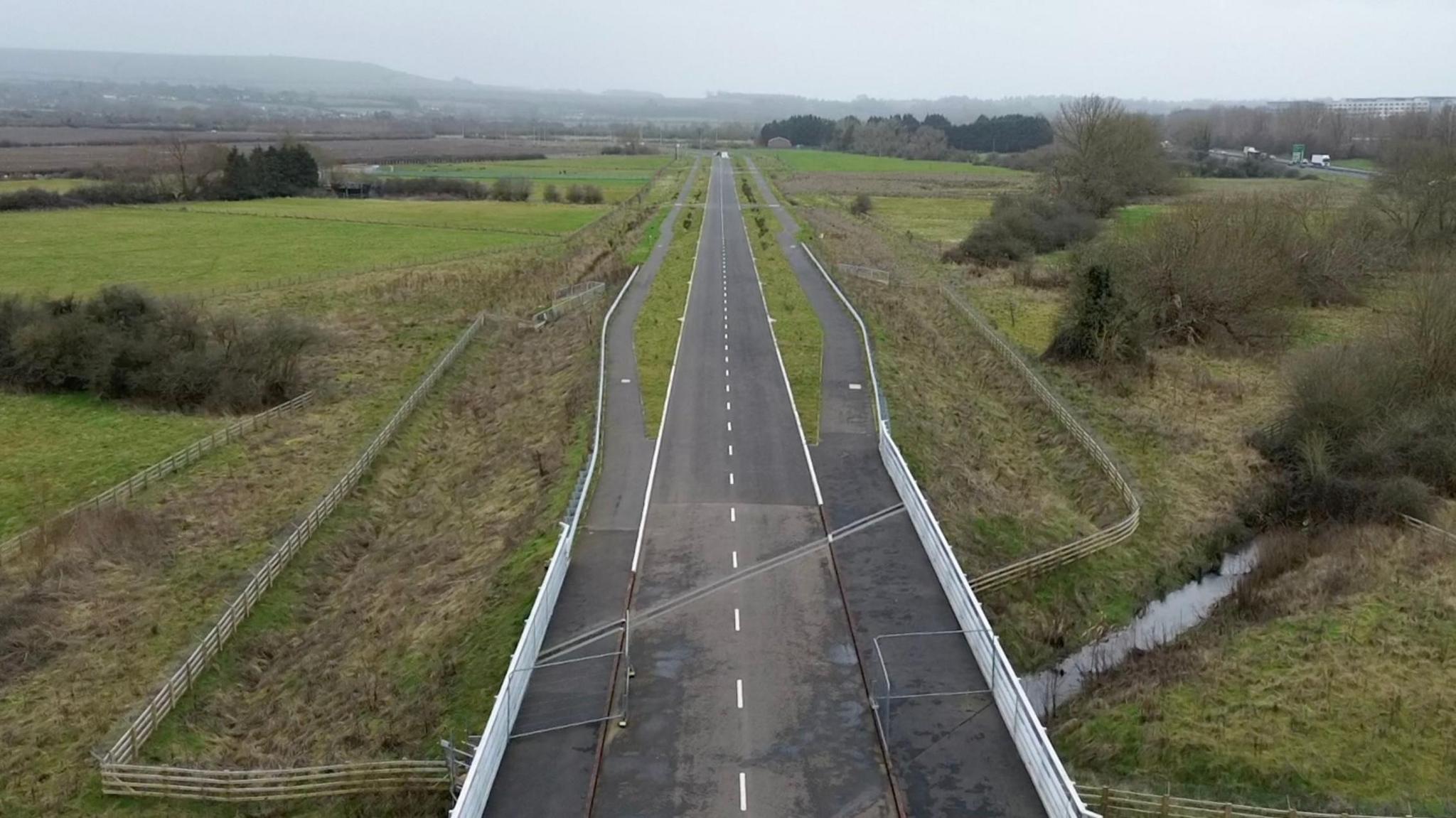 An aerial shot of the unfinished and deserted road near Wanborough. There are fields either side of the Tarmac single-lane carriageway and walking paths immediately next to the road.