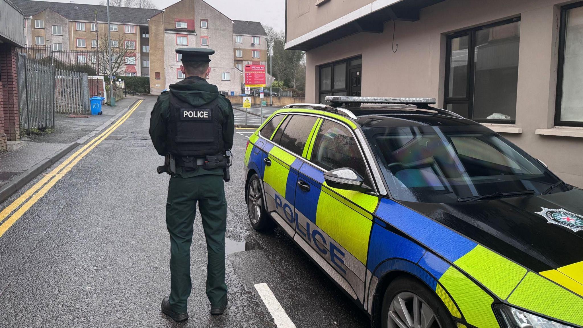 A police officer wearing their uniform stands with their back turned to the camera. In the distance is a small, empty car park. A police car is parked alongside the officer.