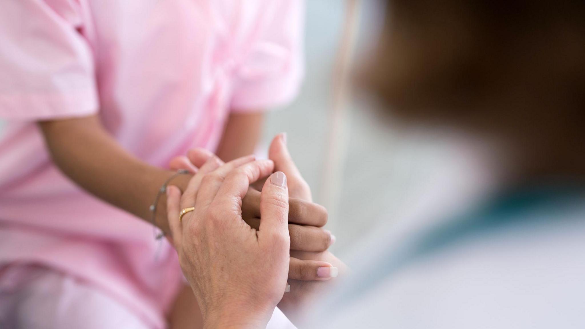 Close up of a nurse in pink scrubs holding a patient's hand.