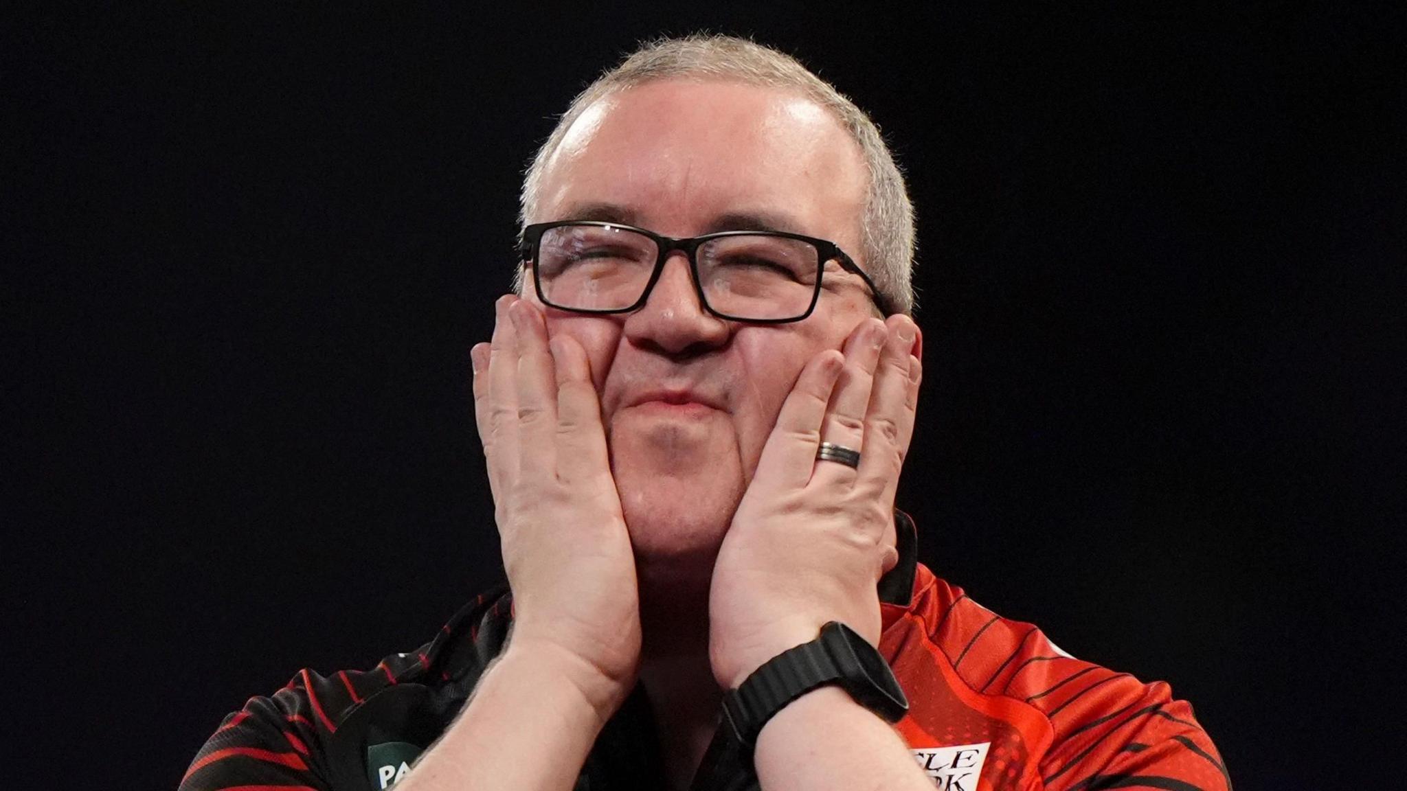 A man with cropped, greying, fair hair wearing black-rimmed glasses and a red and black darts top. His eyes are closed and he is smiling as he holds his hands up to his face. 