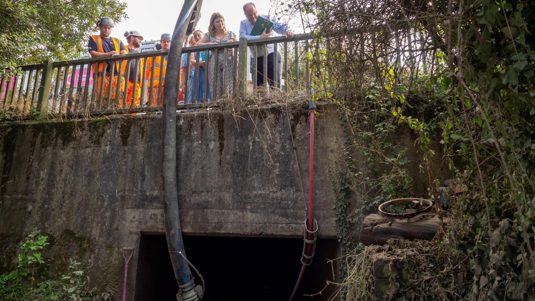 A group of people, men and women peer over a bridge. They are looking into a culvert and pipes to draw debris run into the culvert from a machine on the bridge. Men wearing high vis gear and hard hats are with the group. 