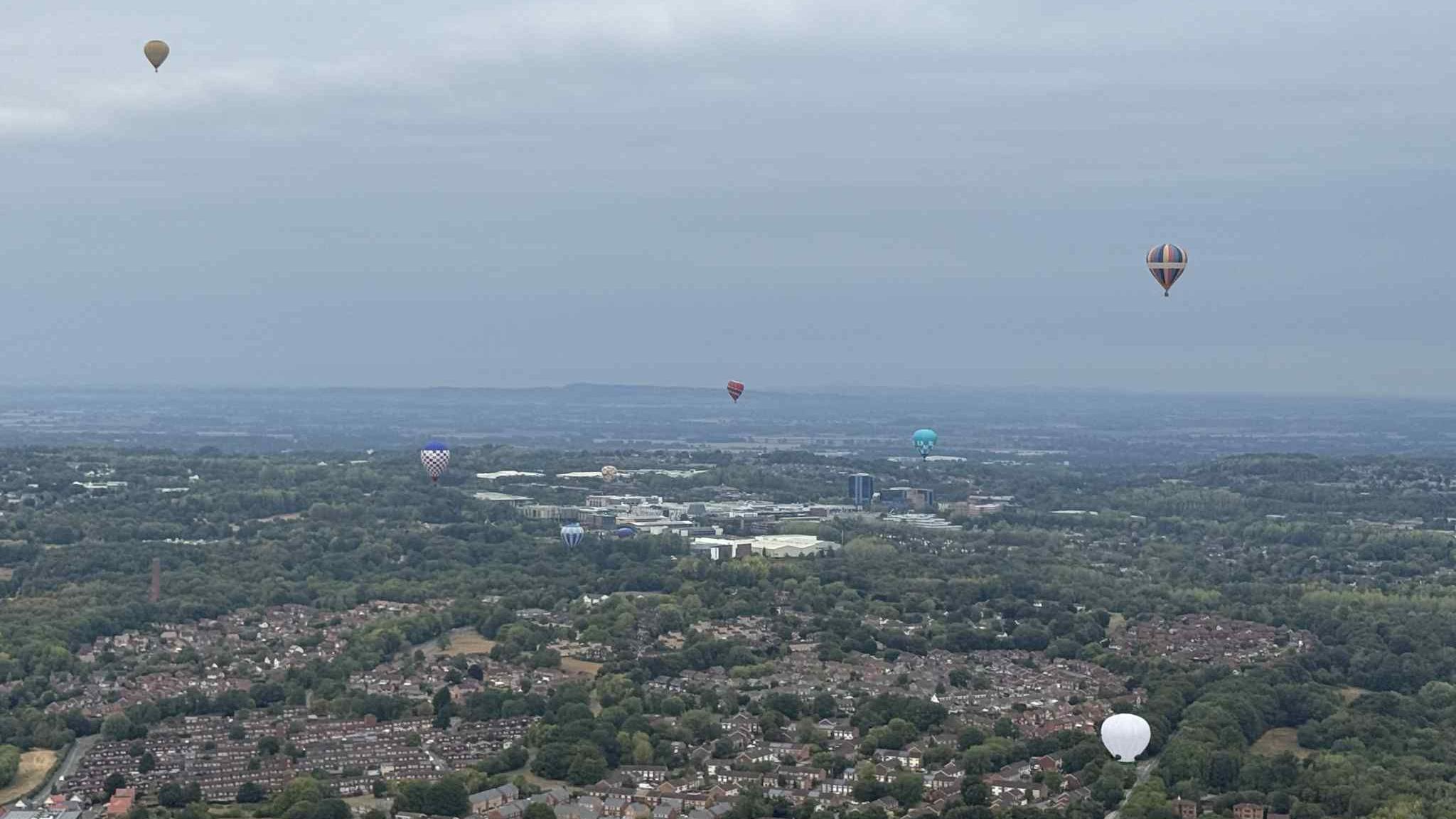 View of multi-coloured hot air balloons in the sky while trees and buildings are below. The skyline looks grey due to the weather.