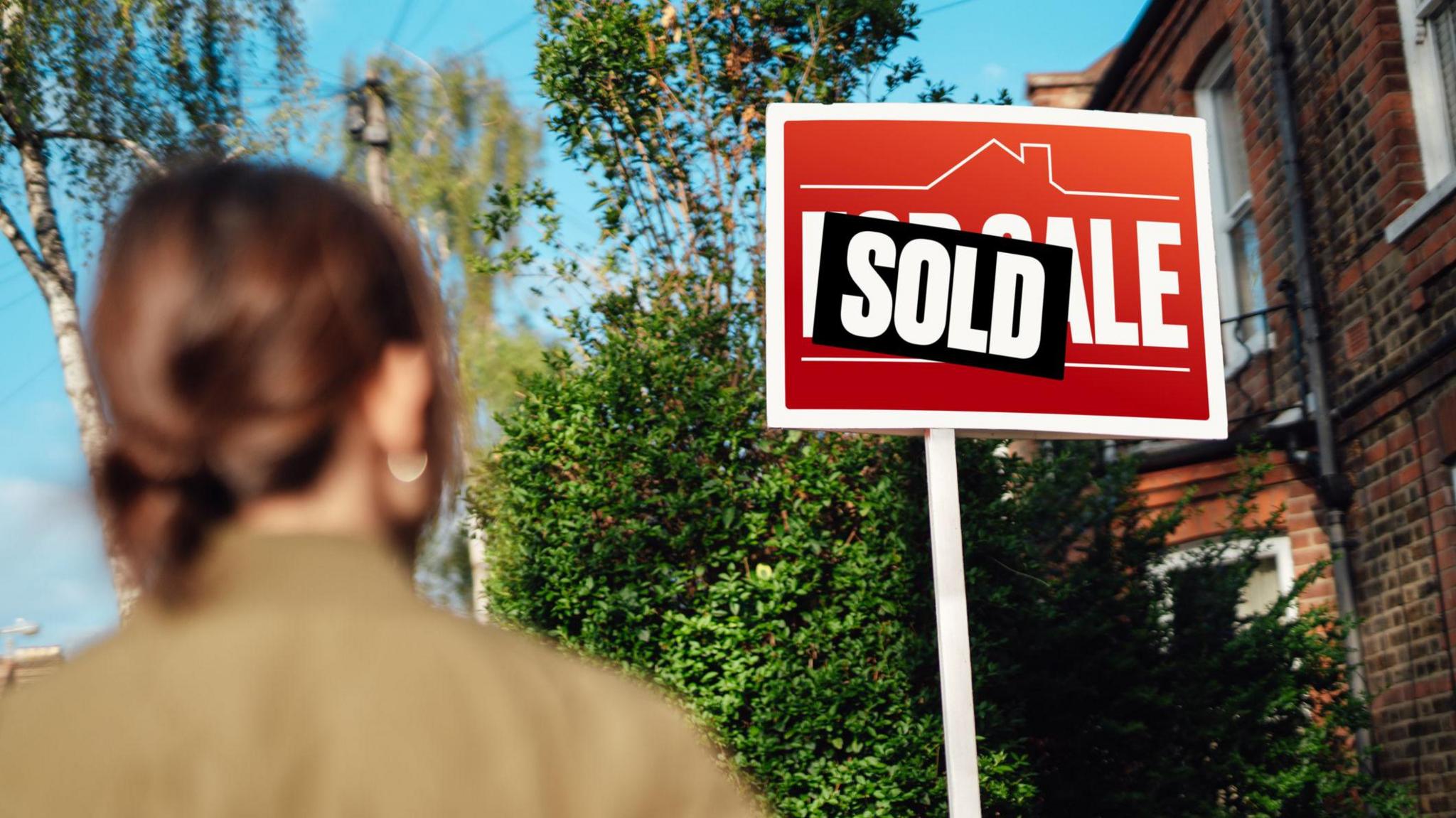 For sale sign covered with a sold sticker, with a woman in the foreground looking at it.