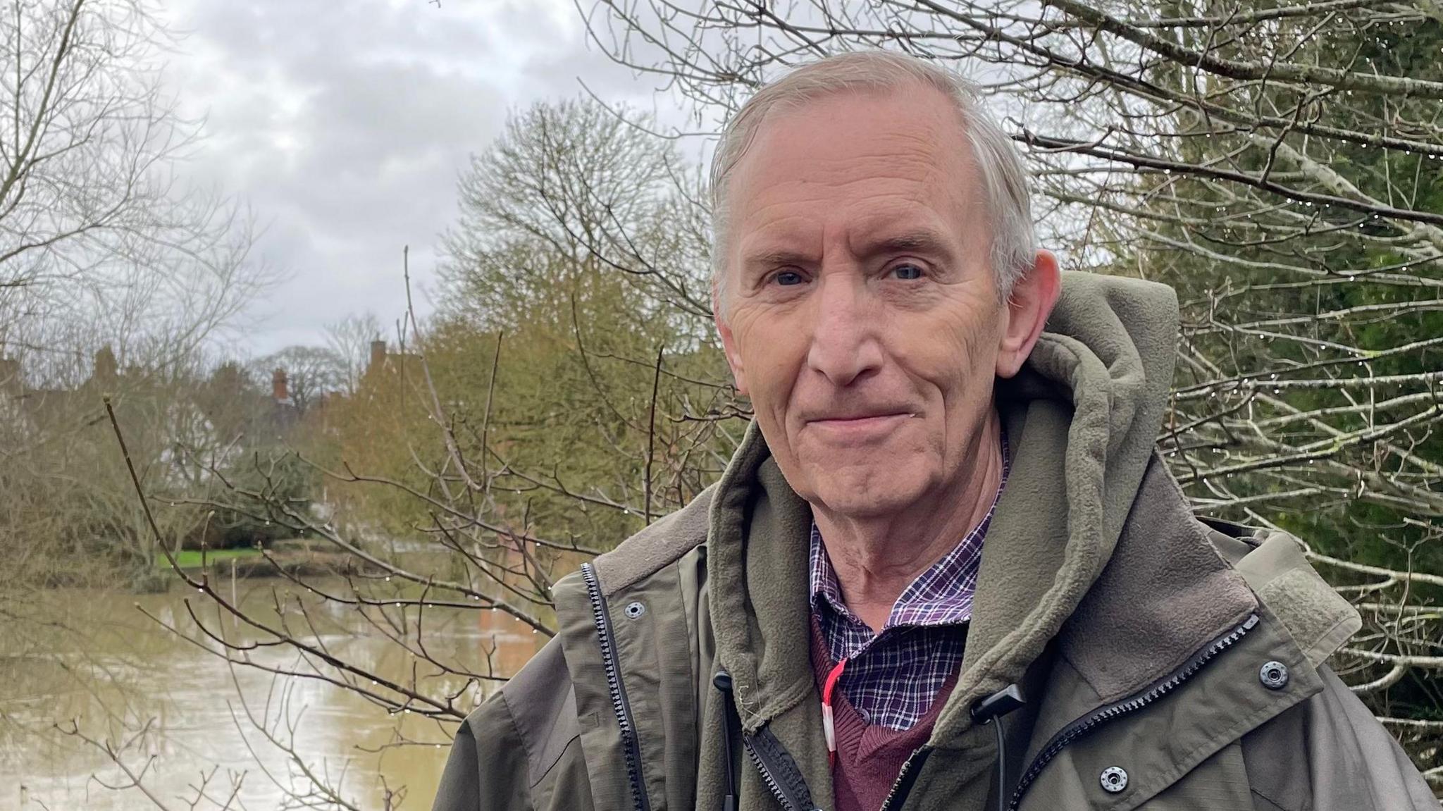 David Adams looking direct to camera and standing in front of a body of water and several trees, most of which have shed their leaves. The body of water is the River Ouzel, which is at flood. Richard has grey hair and is wearing a purple checked shirt, dark red v-neck sweater, khaki hooded top and a heavy, khaki waterproof overcoat. 