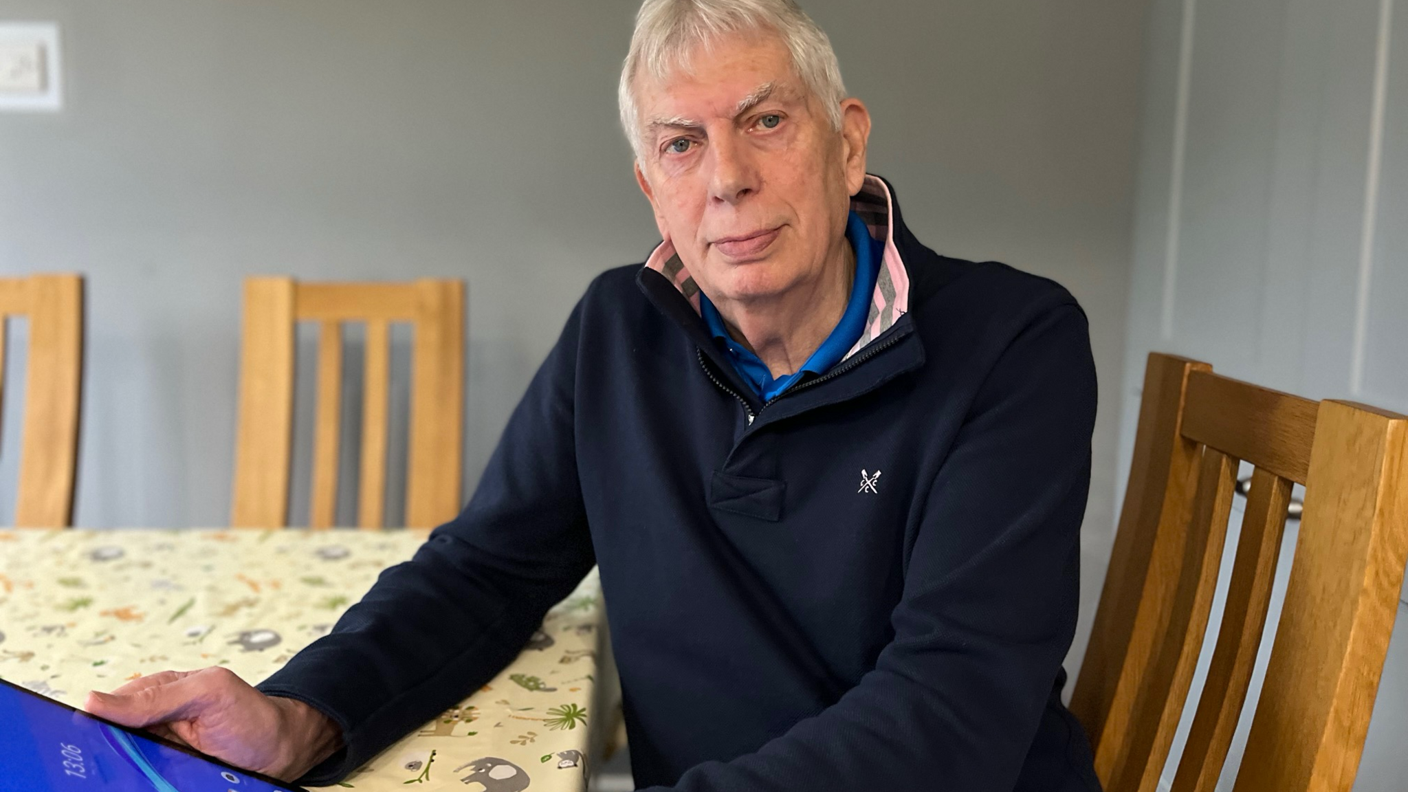 Mike Benbow, a man with white hair and wearing a navy jumper, sits at a kitchen table holding a tablet.