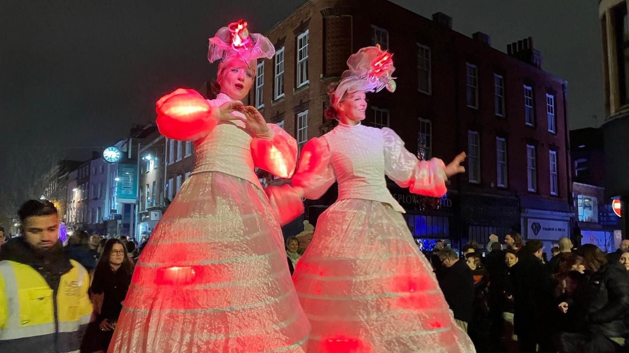 Two women wearing long white dresses which seem to have lights underneath them, and decorative hats adorned with lights, stand in the middle of a crowd of people. They appear to be raised above the people around them, although their feet are not visible.