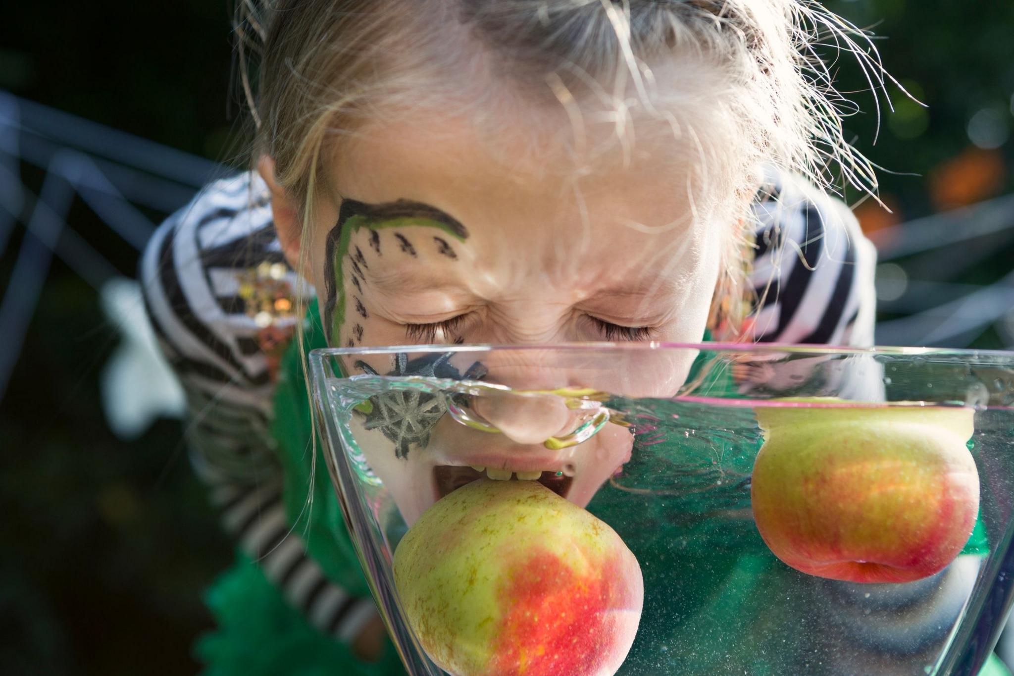 a girl wearing a halloween costume with her eyes closed tries to bite an apple that is floating in a bowl of water