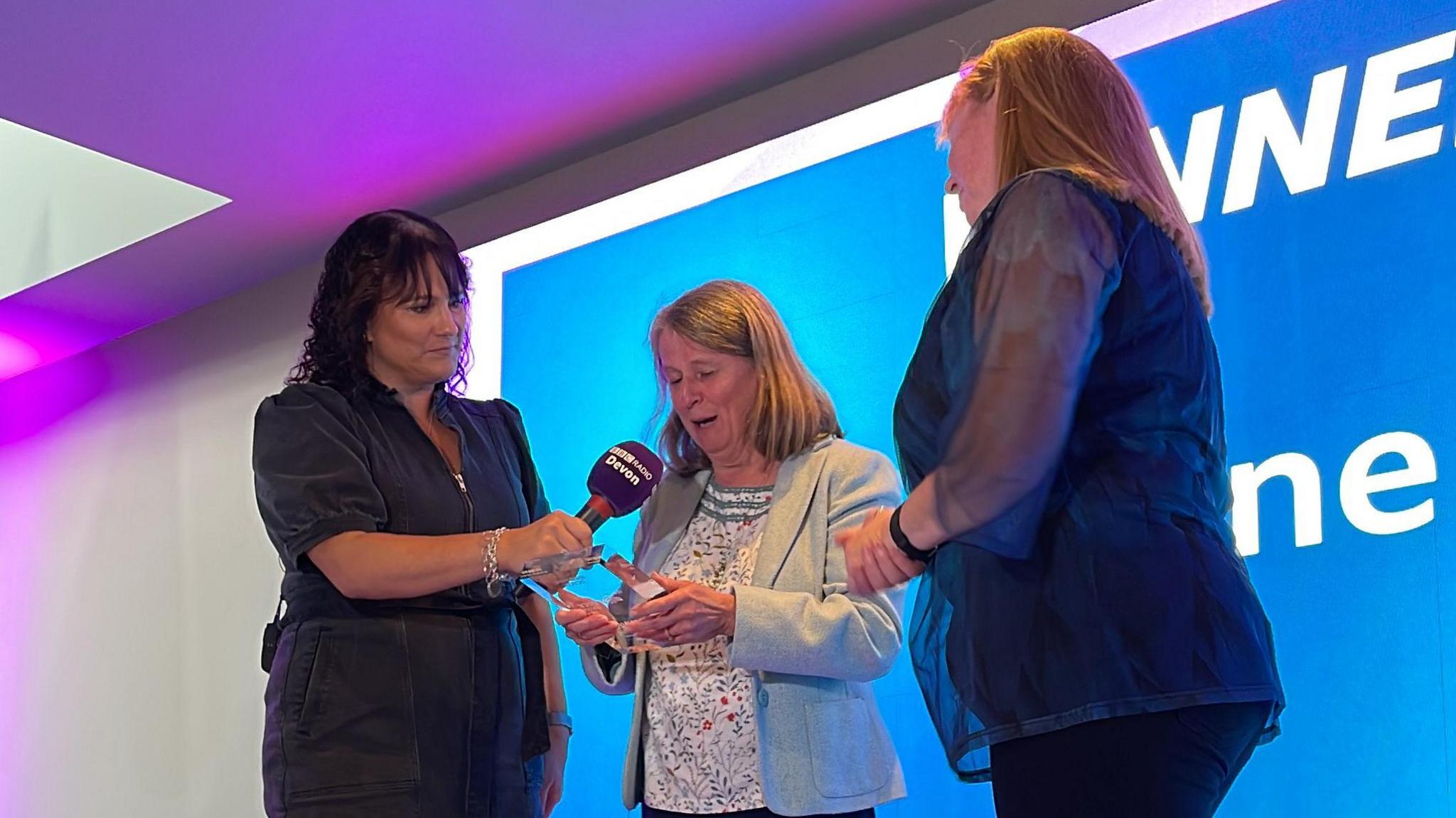 Jane Taylor on stage looking down at the glass award. She is wearing a grey blazer with a speckled top underneath. A woman on the left is holding a purple BBC Radio Devon microphone to Jane and a woman on the right is wearing a dark blue top.