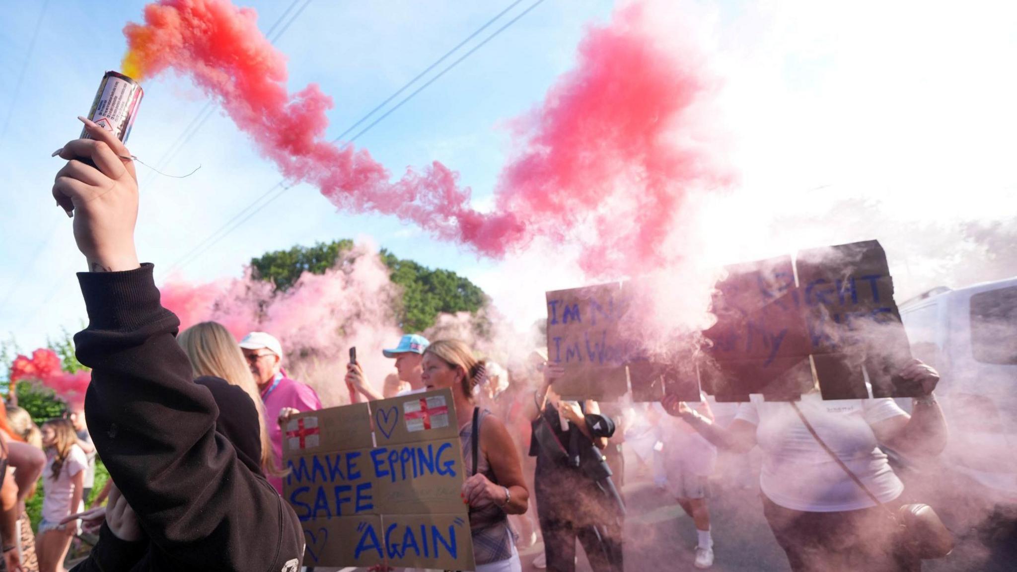 Red smoke pours out of a flare held up by a person, who is part of a larger group marching along a road. Some are holding cardboard placards.