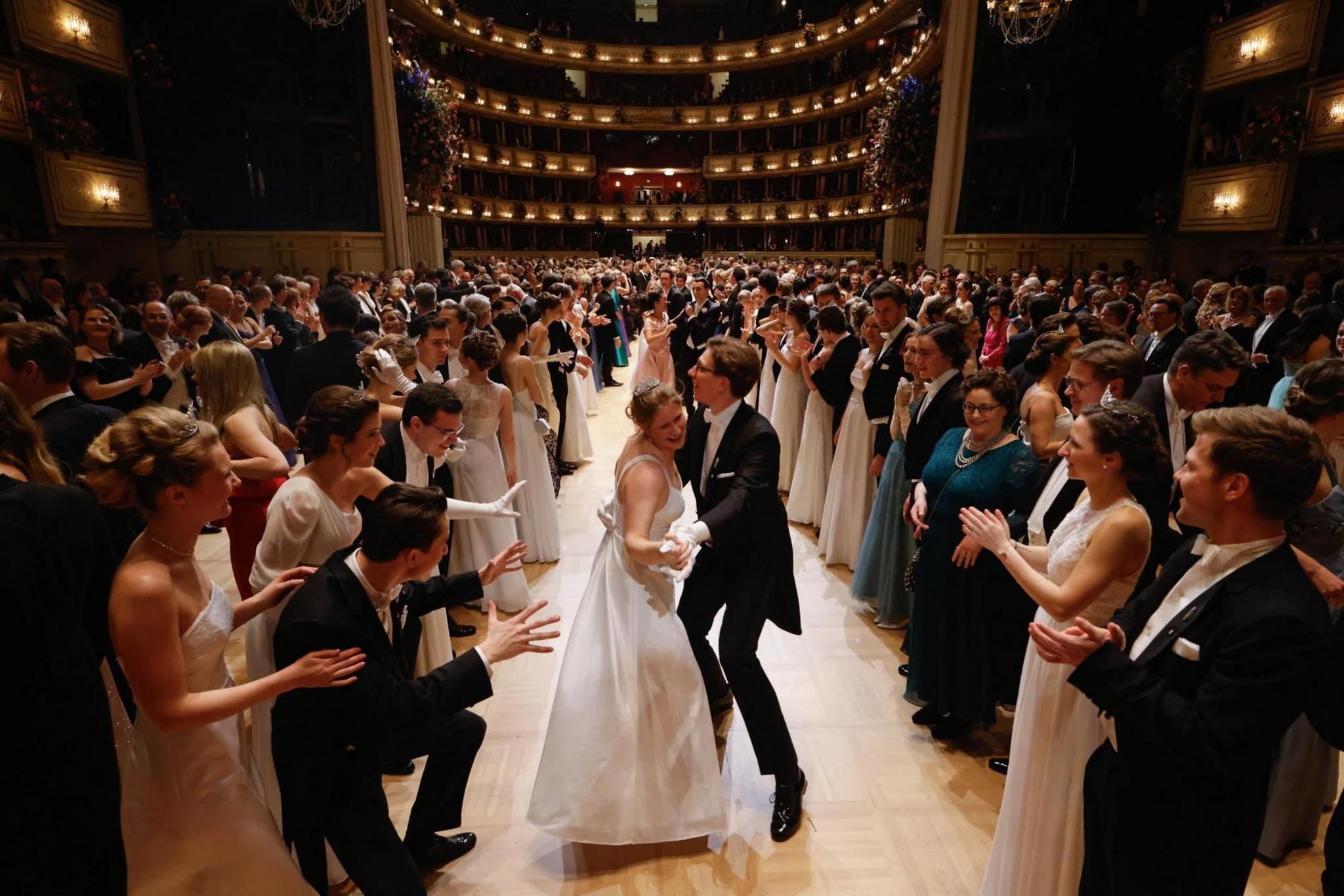 Couples wearing formal attire dance and clap in a grand ballroom. There is a couple in the centre of the image dancing together while others crowd around and watch while clapping