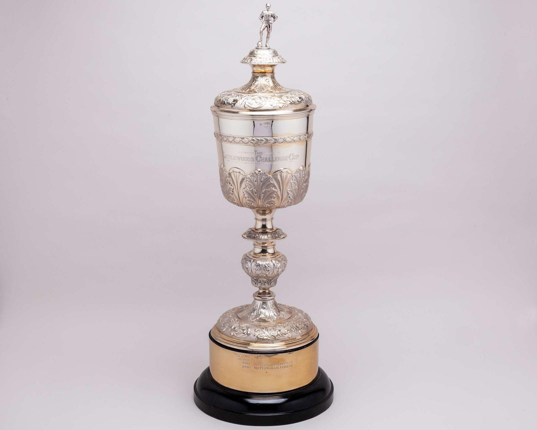 A close-up of a silver trophy inscribed with the words "The Littlewoods Challenge Cup" and a small silver figure of a man with a football on the very top.