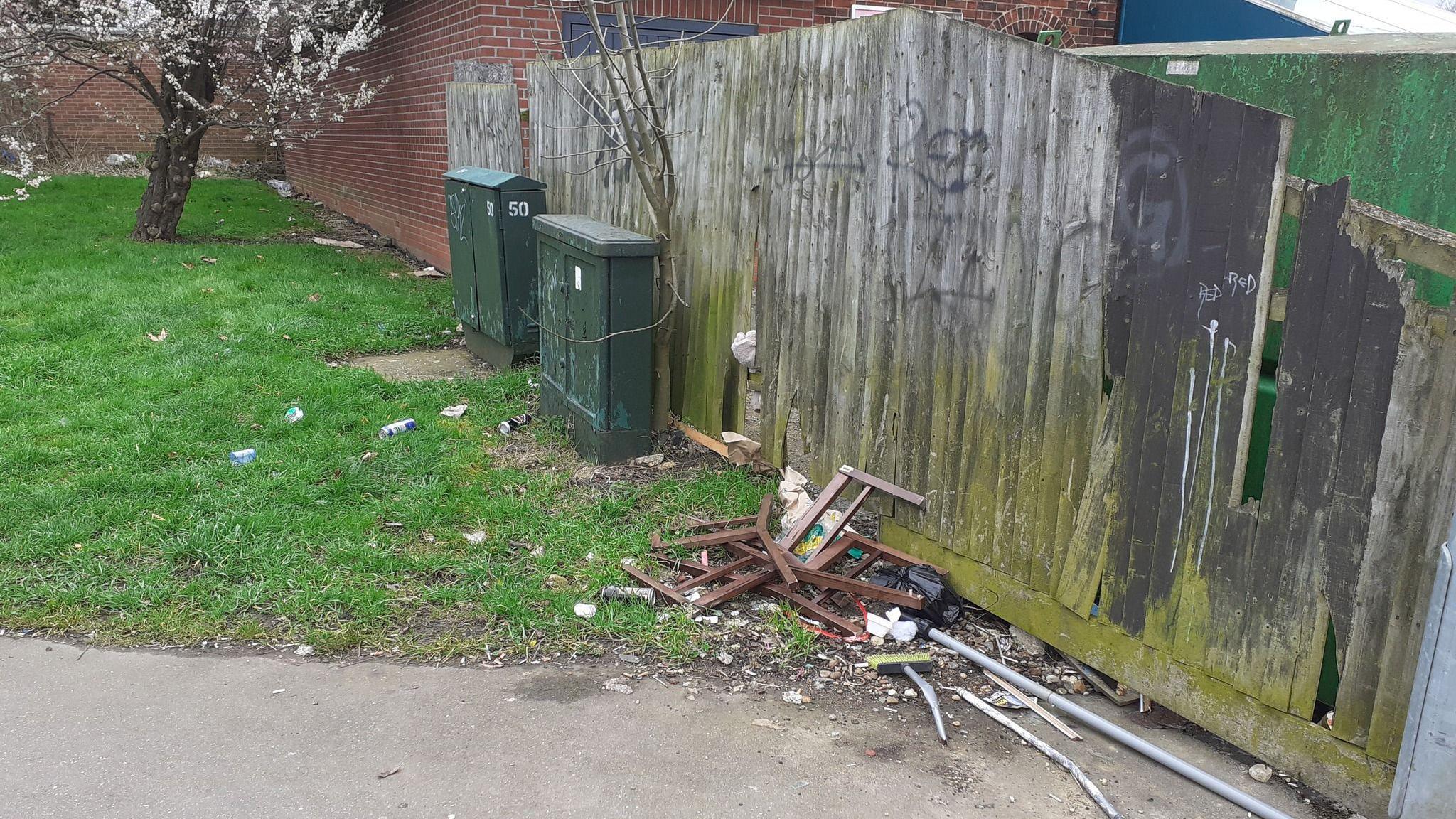 A residential area of Peterborough with a broken wooden fence with graffiti on, there is a grassy verge area, with a tree on but rubbish is strewn around it and near the fence, there is also a smashed wooden chair and some metal silver piping just discarded near it.