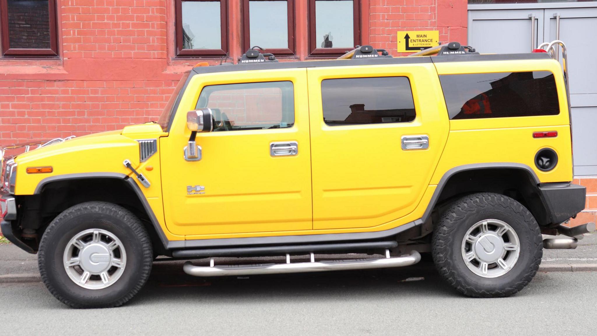 A side view of a yellow Hummer H2 car parked on the street in front of a red brick building