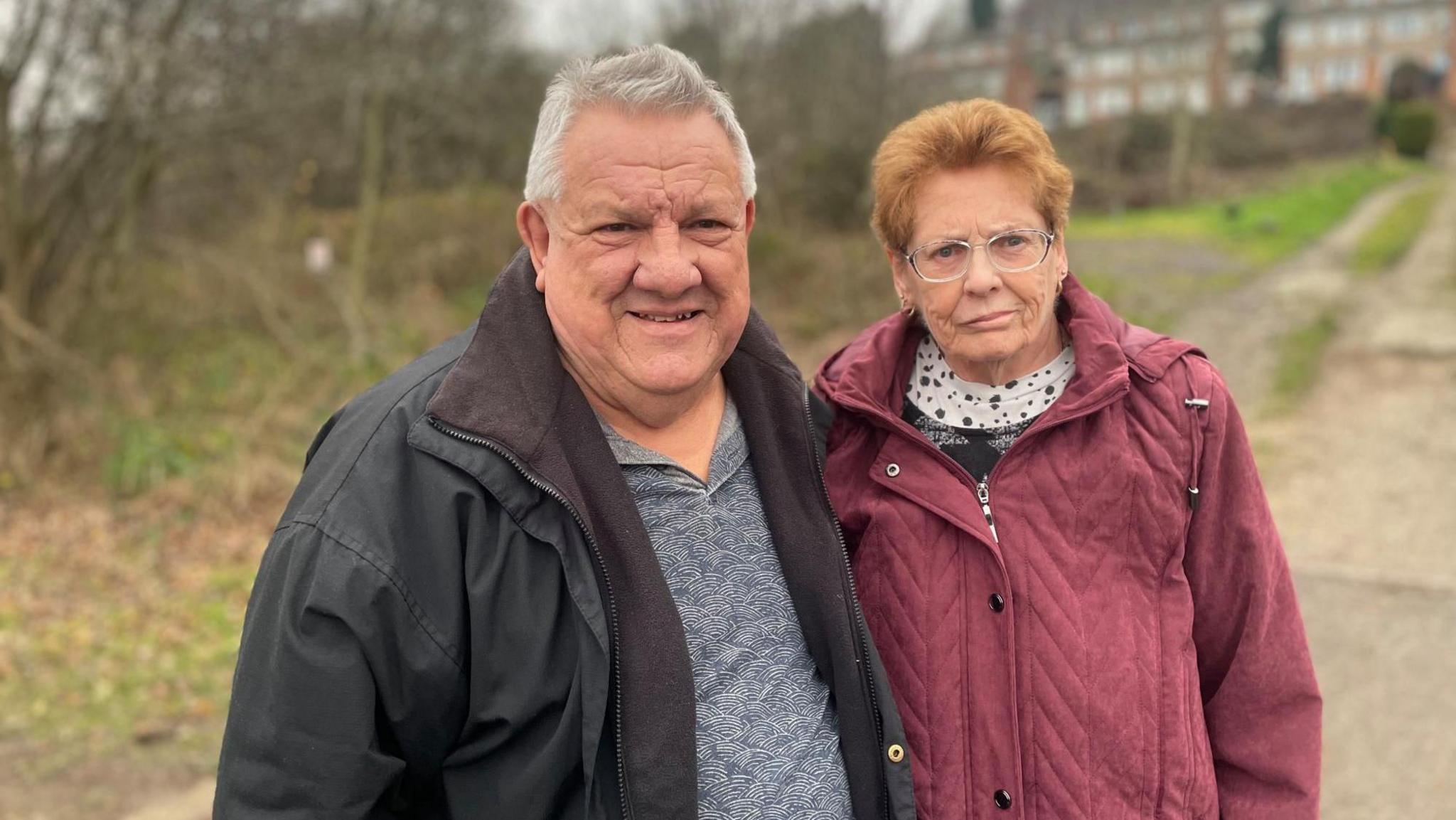 An elderly man in a black coat stands next to an elderly woman in a red coat with a stretch of waste land behind them.