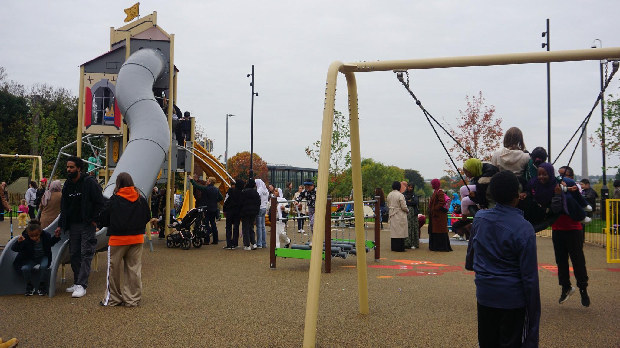 Groups of families and children, in a newly opened play area with children on a swing, coming down a slide, that looks like a castle, other play equipment. The sky is grey and there are trees in the distance.