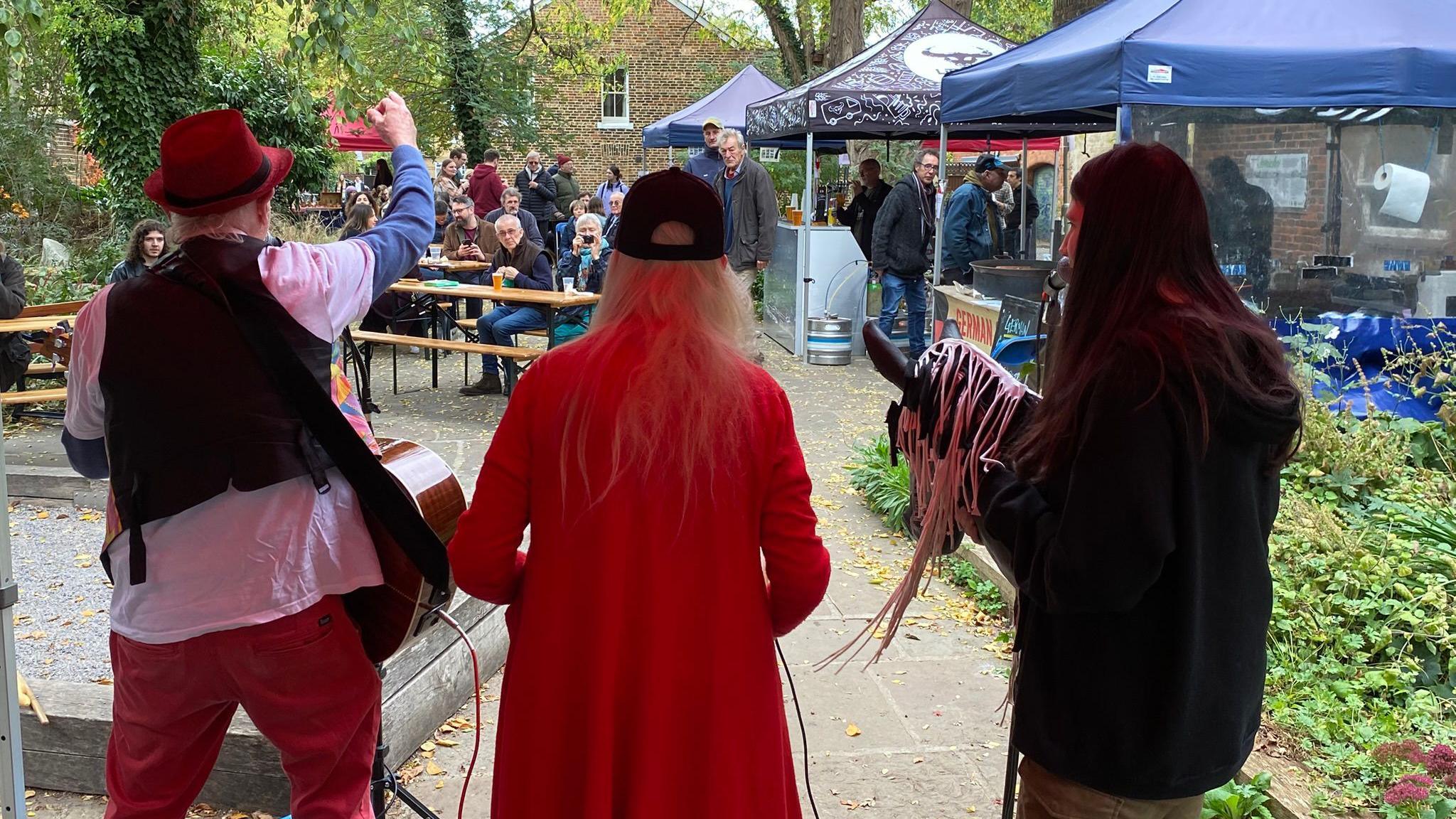 A shot taken from behind a band performing at Jericho Oktoberfest on 17 October. The crowd is looking at the singer and musicians. One of them is holding a hobby horse as part of the performance. It's cloudy.
