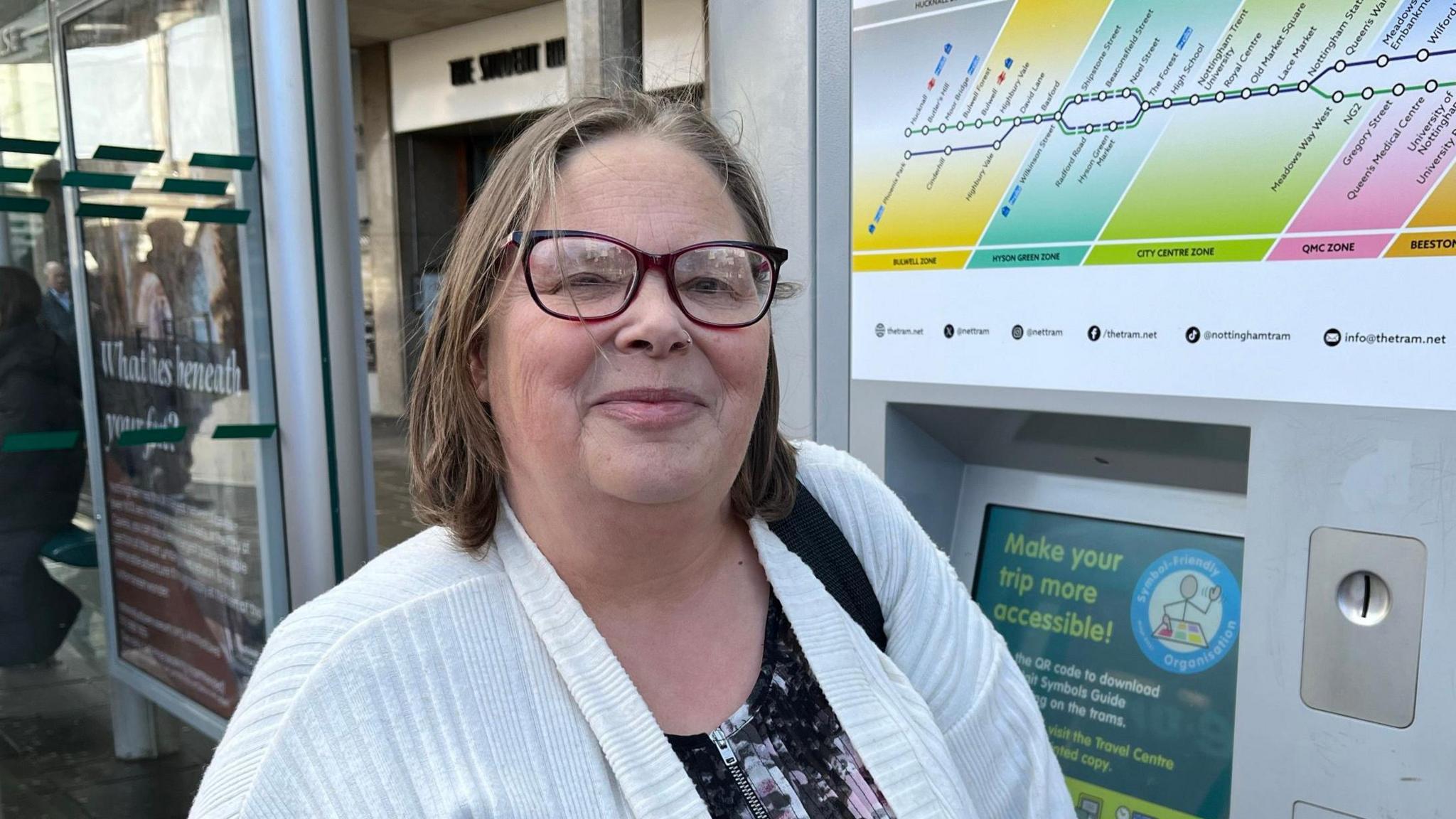 Sarah Peachey, a middle-aged white woman with short brown hair and dark-red-framed glasses. She is standing in front of a tram ticket machine in Nottingham.