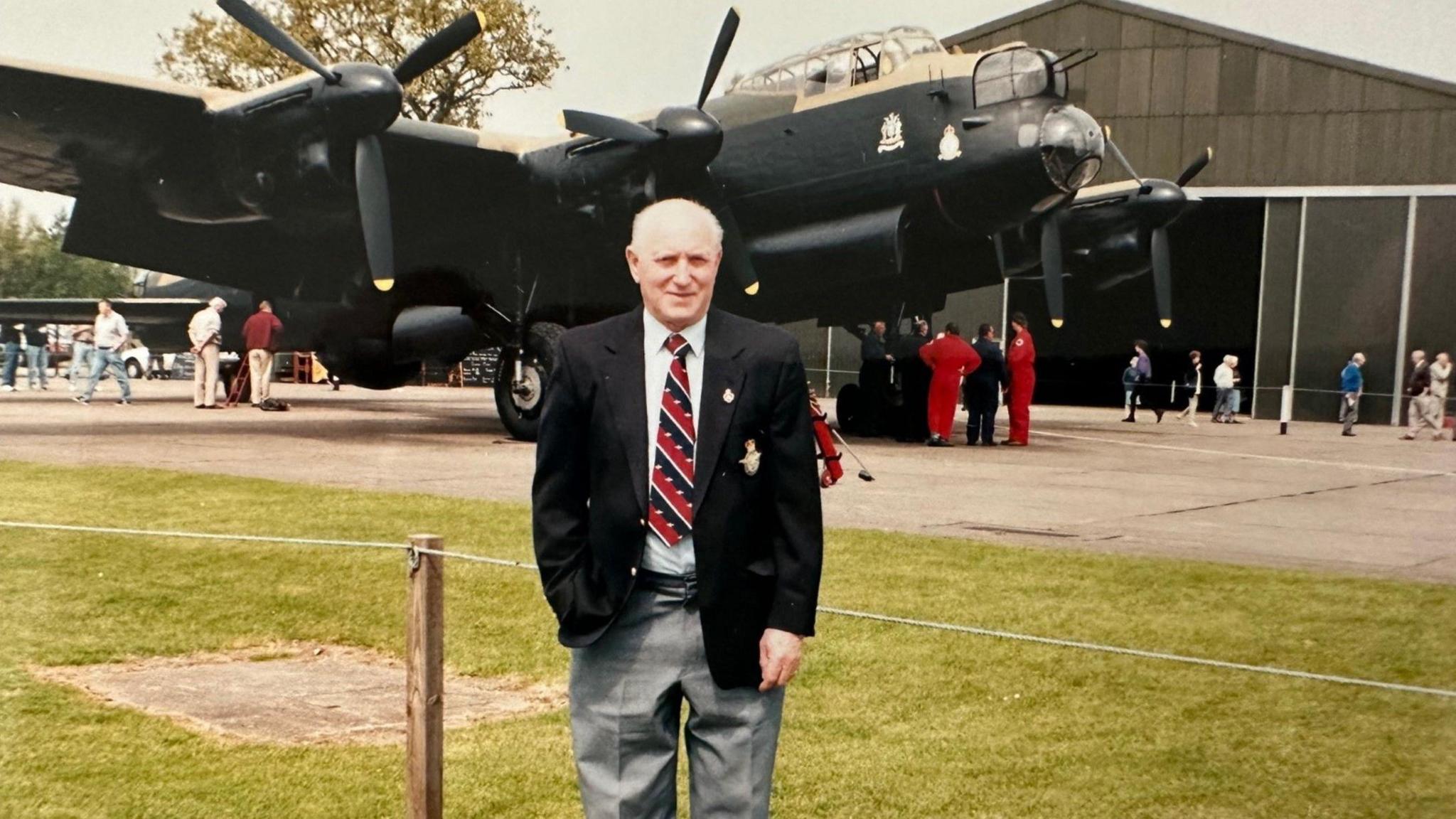 Ivor Taylor standing in front of a Lancaster bomber, which is on display at an airfield. He is wearing grey trousers, a white shirt, black blazer and a red and black striped tie.