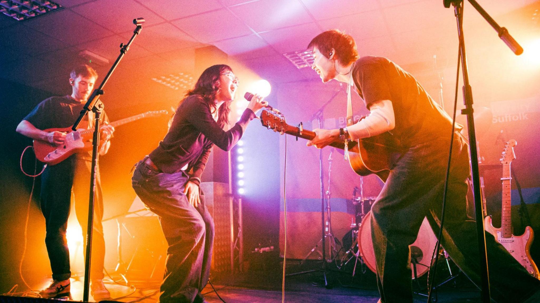 Young female and male lean into each other as they sing on a stage. The woman wears glasses and holds a microphone and the man plays a guitar. Another guitarist is playing on stage too.