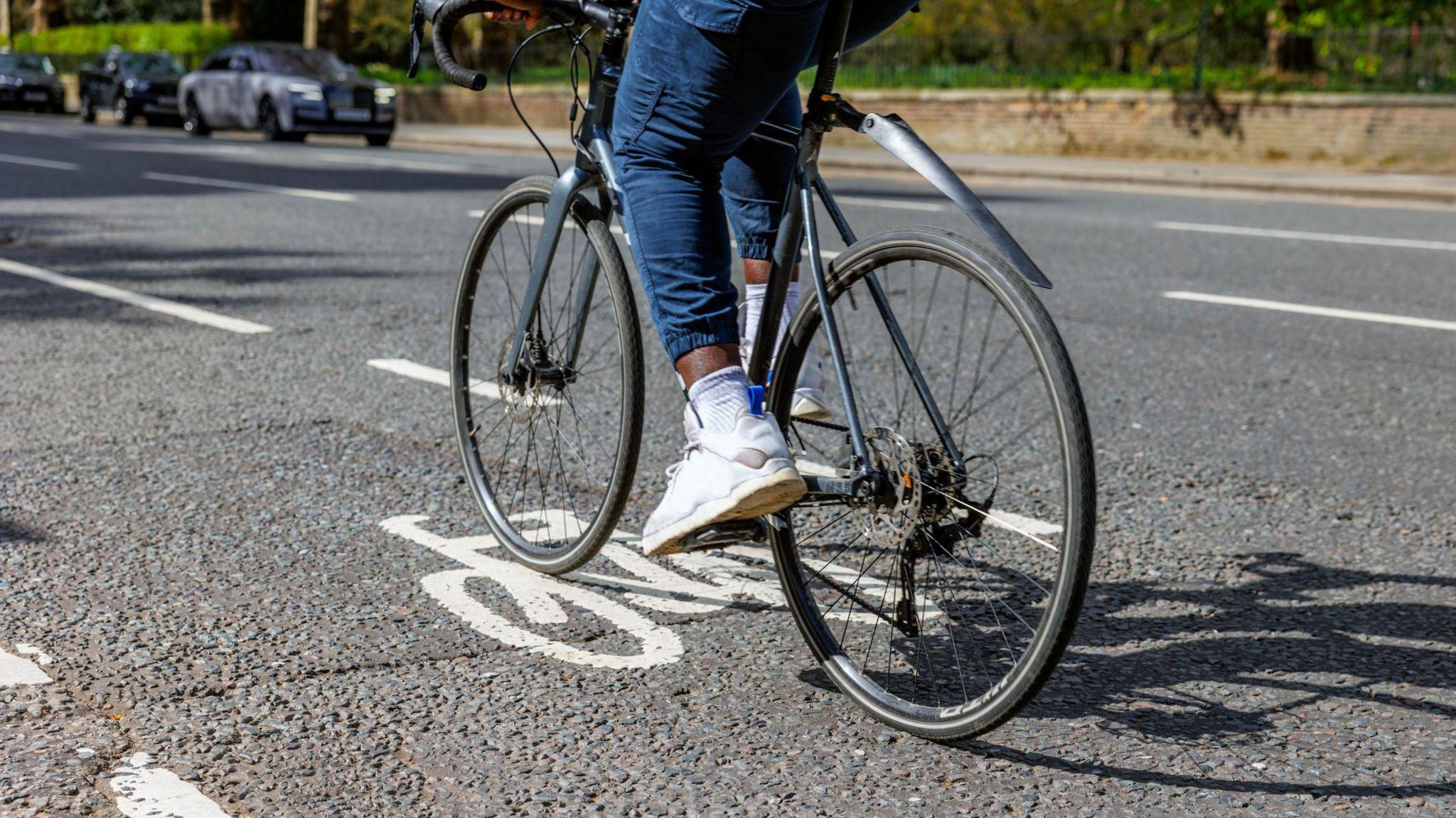 A stock image of a person cycling in a cycle lane. The person is wearing cropped jeans with an elasticated ankle, white trainers and white socks. They are cycling on a section of road, in the background cars can be seen travelling in the opposite direction and behind them is a section of pavement, lined by a short brick walls with hedging.