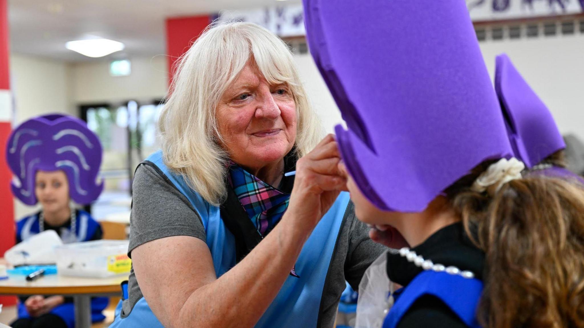 A woman with grey hair with a tartan scarf round her neck applies make-up to a participant in the Dumfries street parade