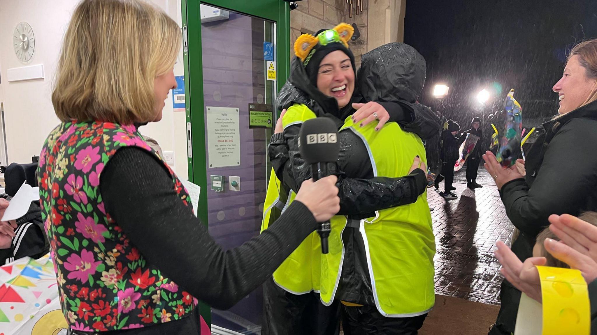 A smiling Amy Garcia and Lindsey Burrow embrace each other after completing their walk. Both women are wearing high-vis vests and black jackets with their hoods up and black trousers.