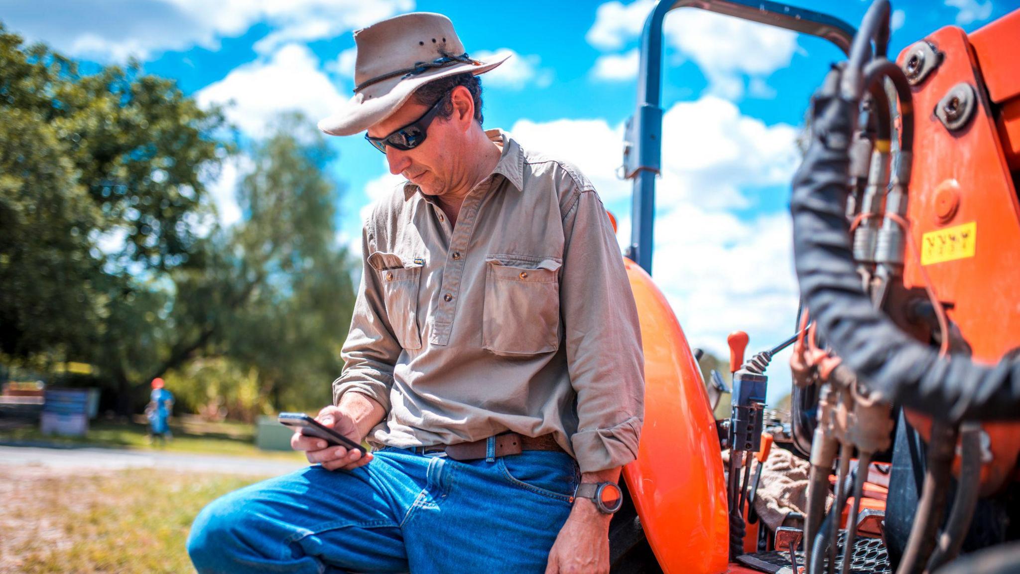 A farmer wearing a hat, sunglasses, jeans and shirt leans on a tractor and looks at a smartphone.
