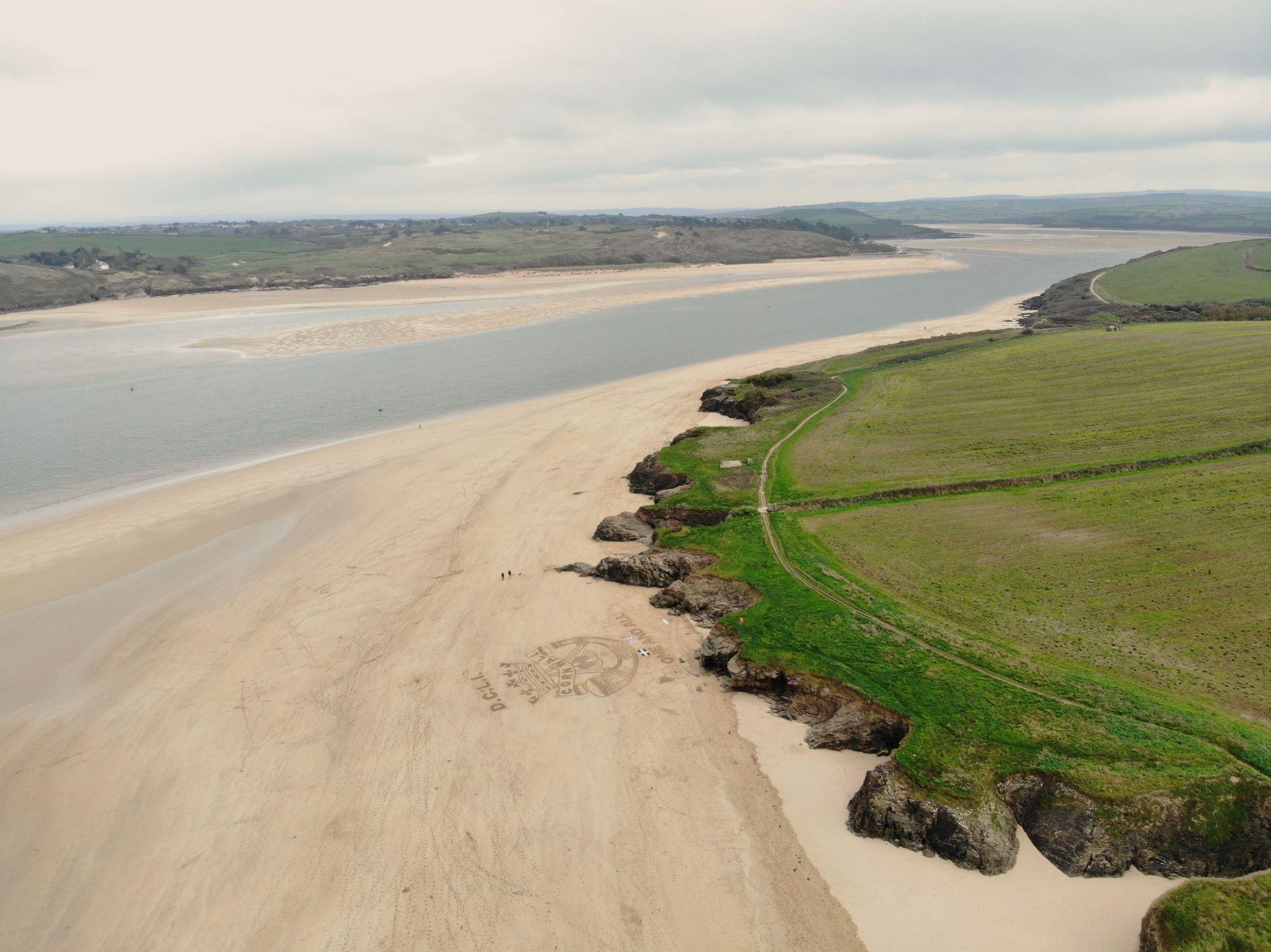 A aerial photo showing the drawing sitting at the base of a cliff that is topped by grass and continues to stretch away into the distance. The tide is out and beyond the sea another stretch of land can be seen. 