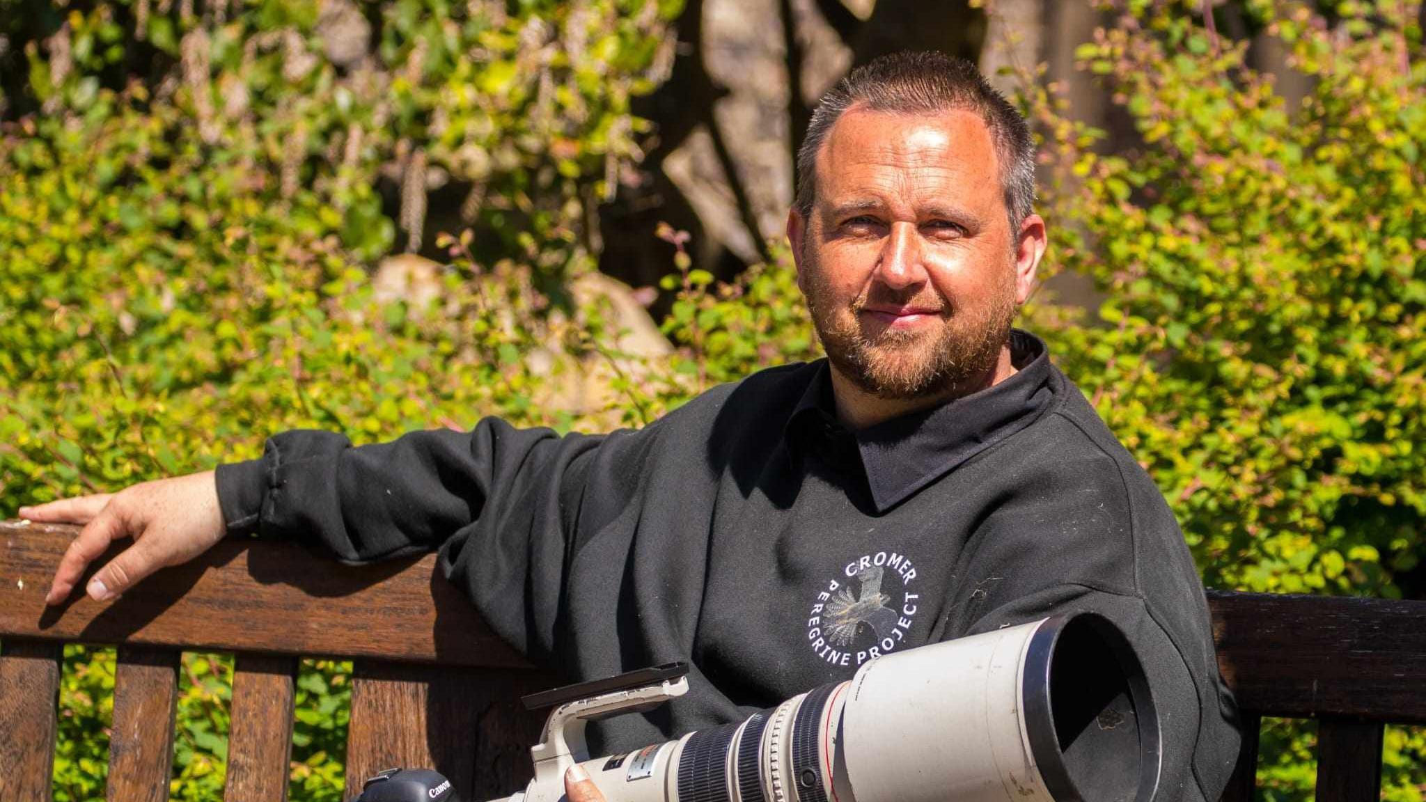 Chris Skipper - A man sitting on a bench with short hair, holding a professional camera with a long lens