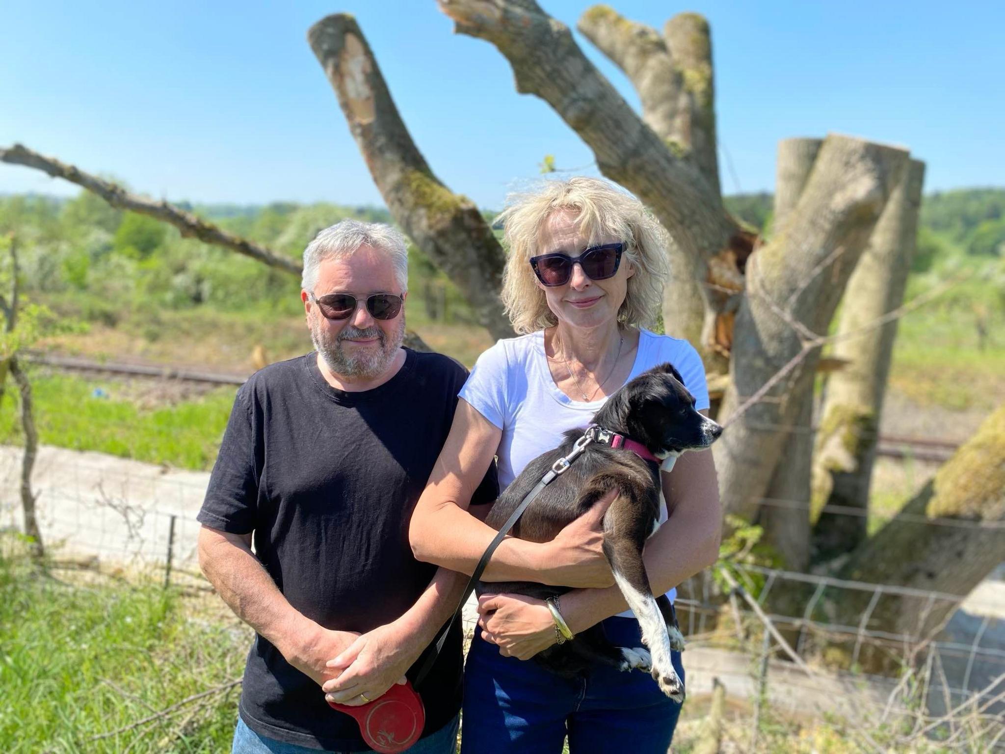 Andy on the left is wearing a black T-shirt and Sarah on the right is wearing a white T-shirt. She is holding a small dog and they are stood in front of a felled tree.