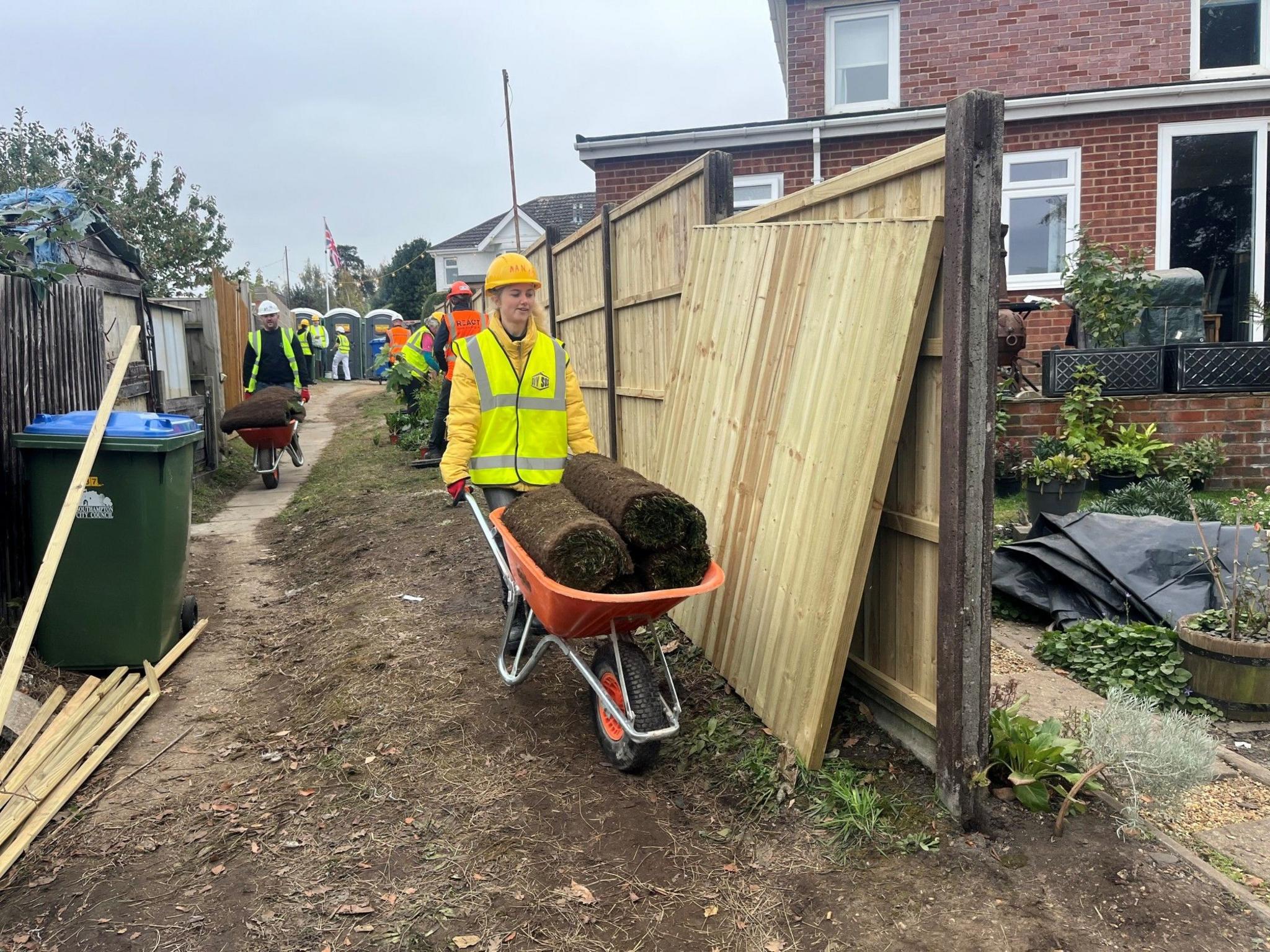 A woman wheeling an orange wheelbarrow full of rolls of turf. She walking to a gap between a wooden fence wearing a florescent jacket and hard hat.