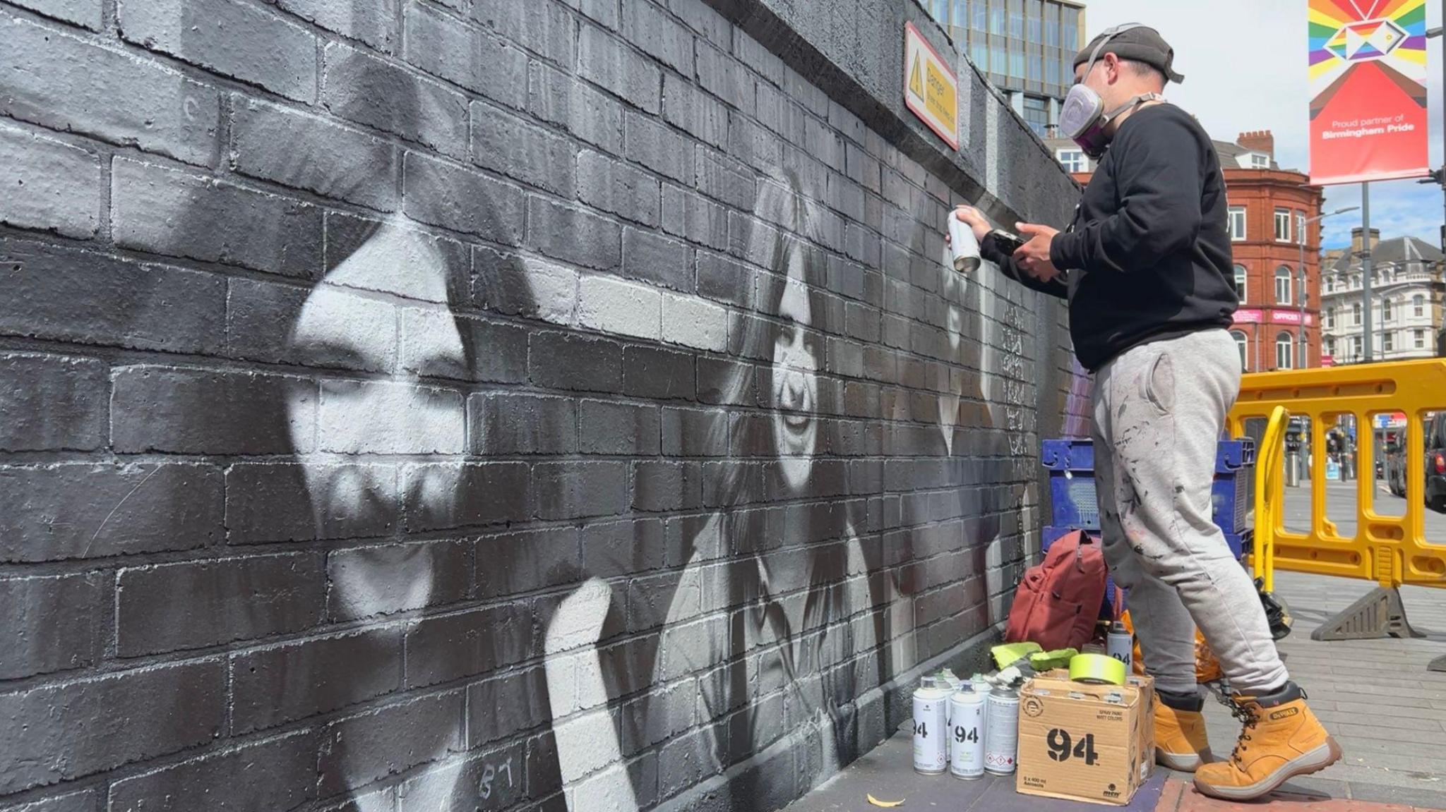 Mr Murals next to his murals in Birmingham. He is working on a 120ft (36.5m) mural depicting all four original members of Black Sabbath. He has a purple mask hanging around his neck. There is also a trolley with paint cans nearby.