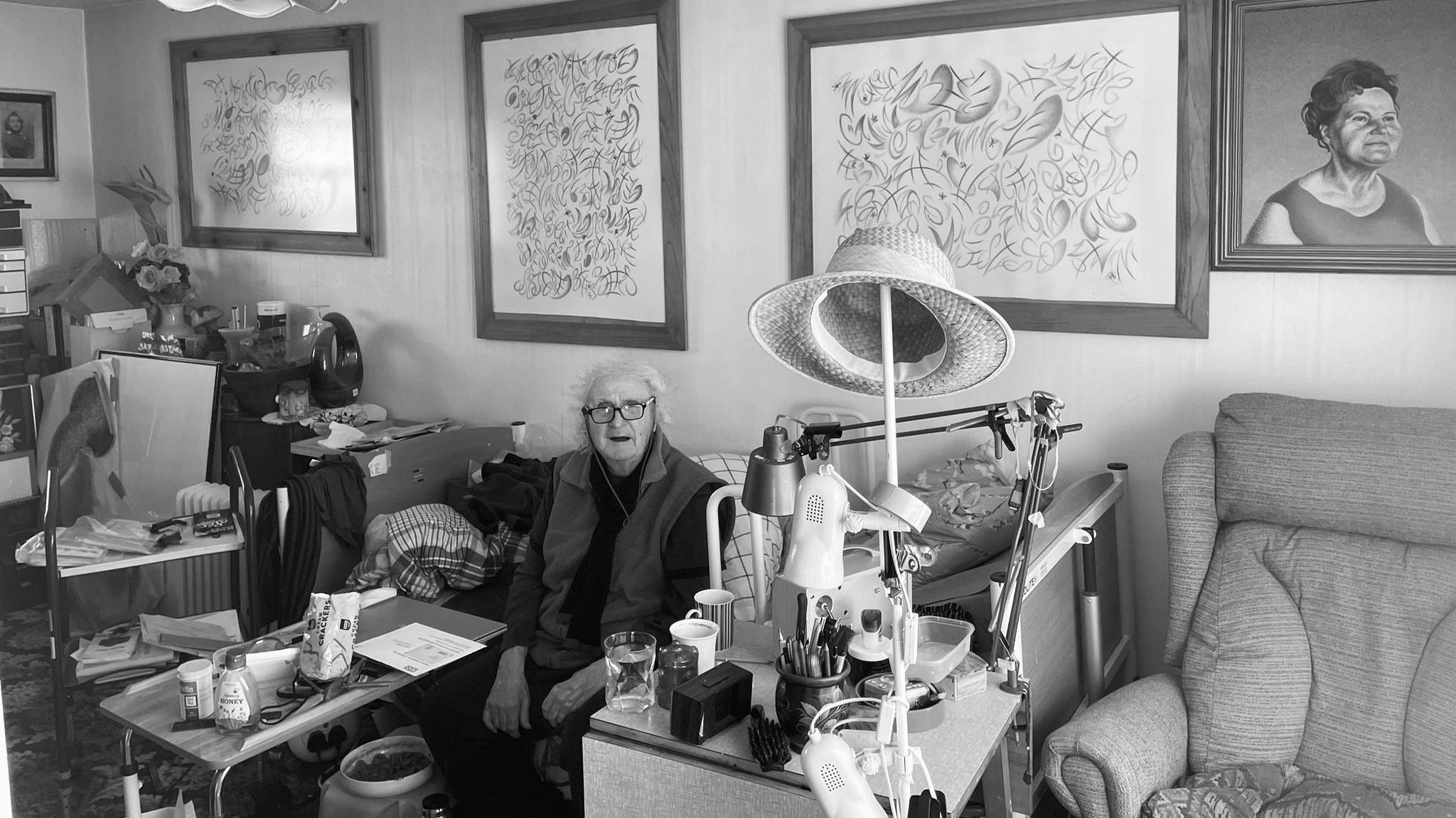 Black & white photo with Henry Orlik in his 70s in a living room with framed paintings behind him as she sits down, with a sofa to the right
